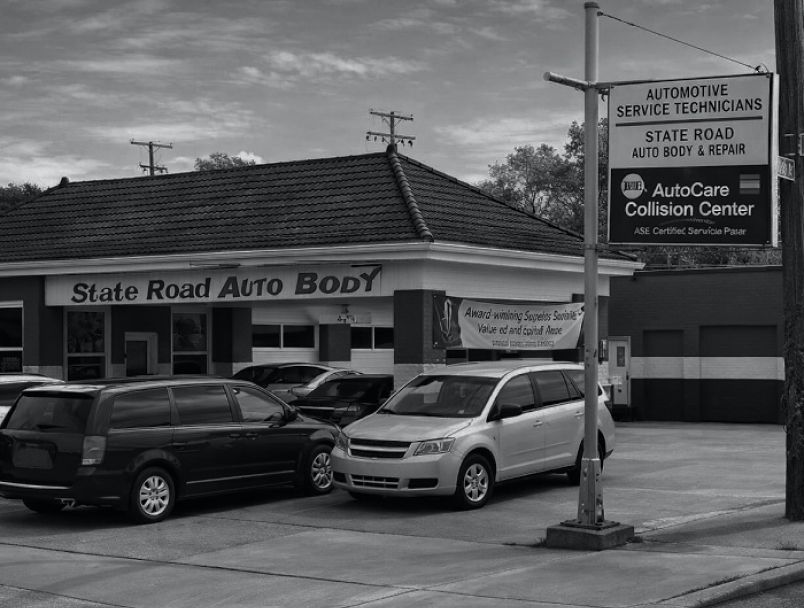 Black and white shot of State Road Auto Body with a service sign, parked cars, and a repair shop building. | State Road Auto Body & Repair