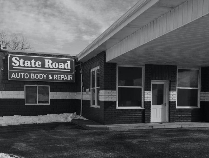 A brick commercial building with a sign reading "State Road AUTO BODY & REPAIR" under a partially covered driveway. | State Road Auto Body & Repair