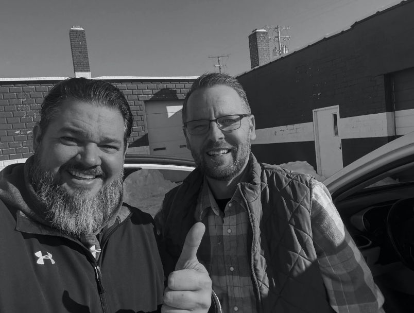 Two men smiling outdoors in front of a building, one with a beard and the other giving a thumbs-up. | State Road Auto Body & Repair