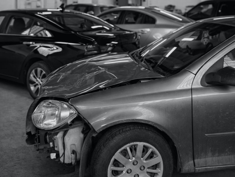 A damaged car with a crushed hood and missing front bumper in a garage with other vehicles. | State Road Auto Body & Repair