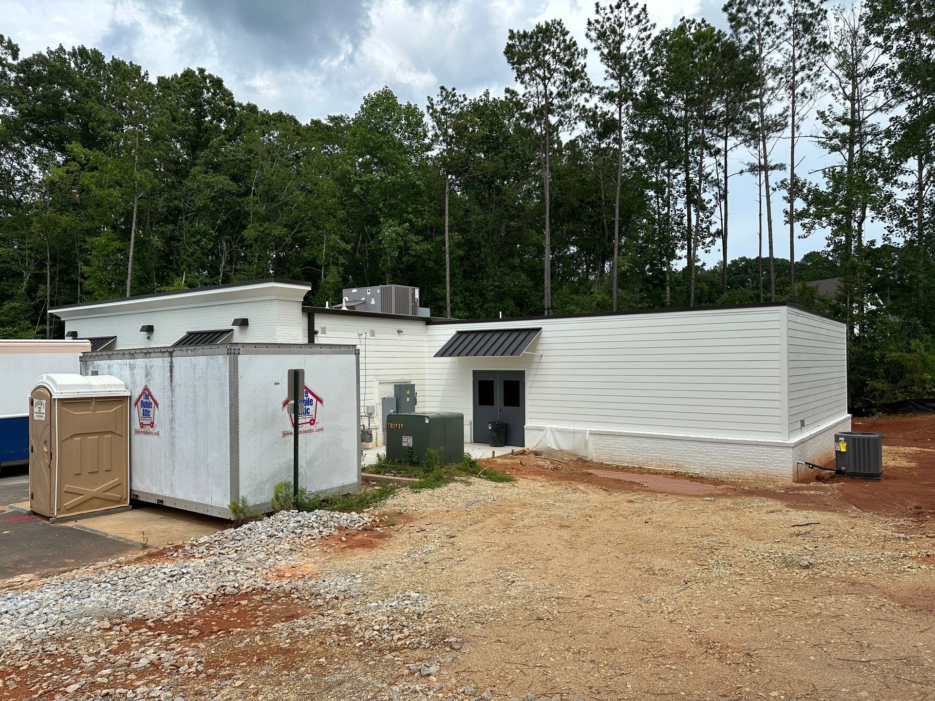 A large white building is sitting in the middle of a dirt field surrounded by trees.