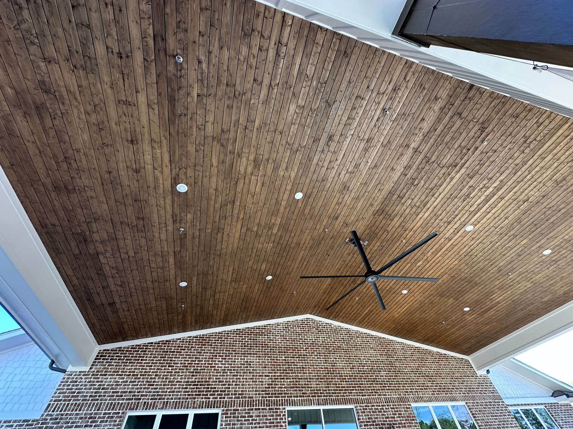 A covered outdoor patio featuring a wooden plank ceiling, a large ceiling fan, and recessed lighting above a brick wall.