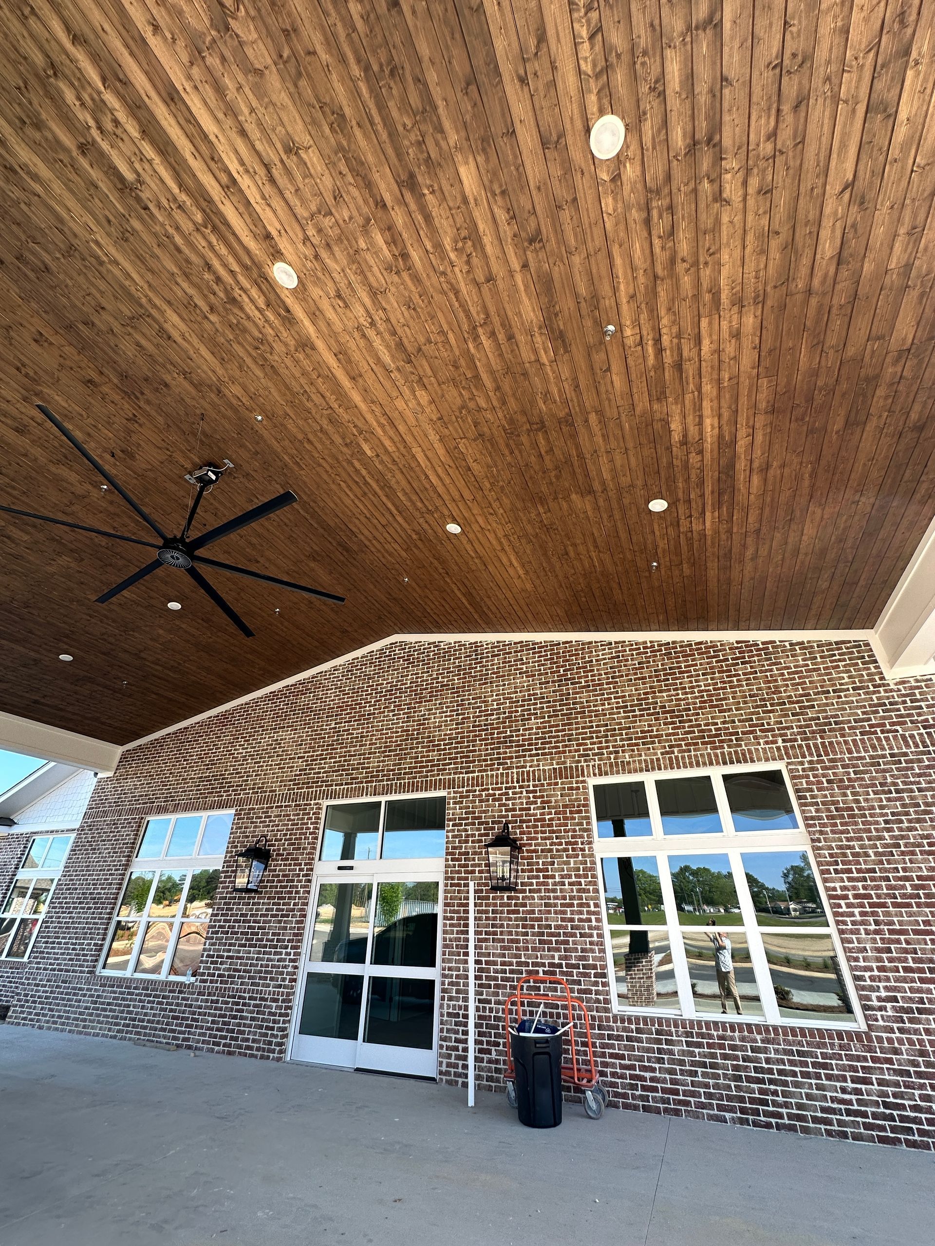 Exterior of a building featuring a dark wood-paneled covered porch, brick walls, glass doors, and windows under a blue sky.