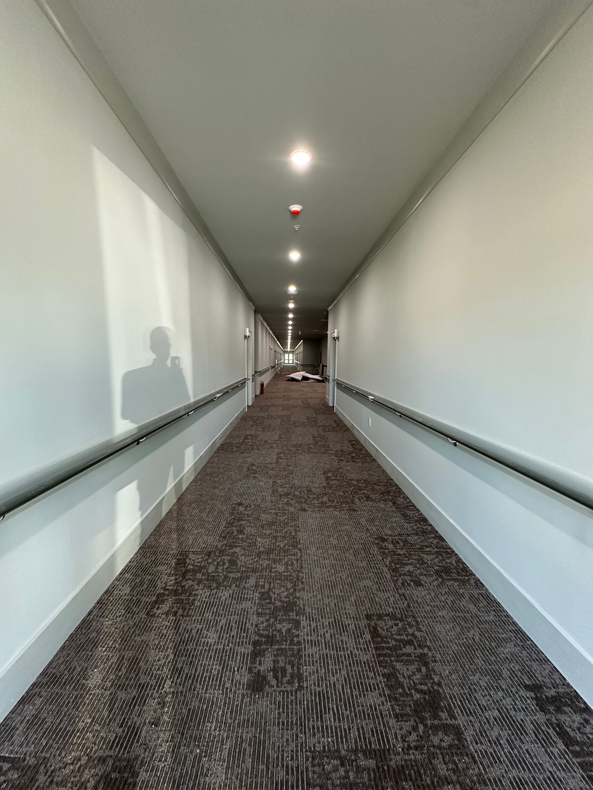 A long, narrow hotel corridor with a dark patterned carpet, white walls, handrails, and recessed ceiling lighting.