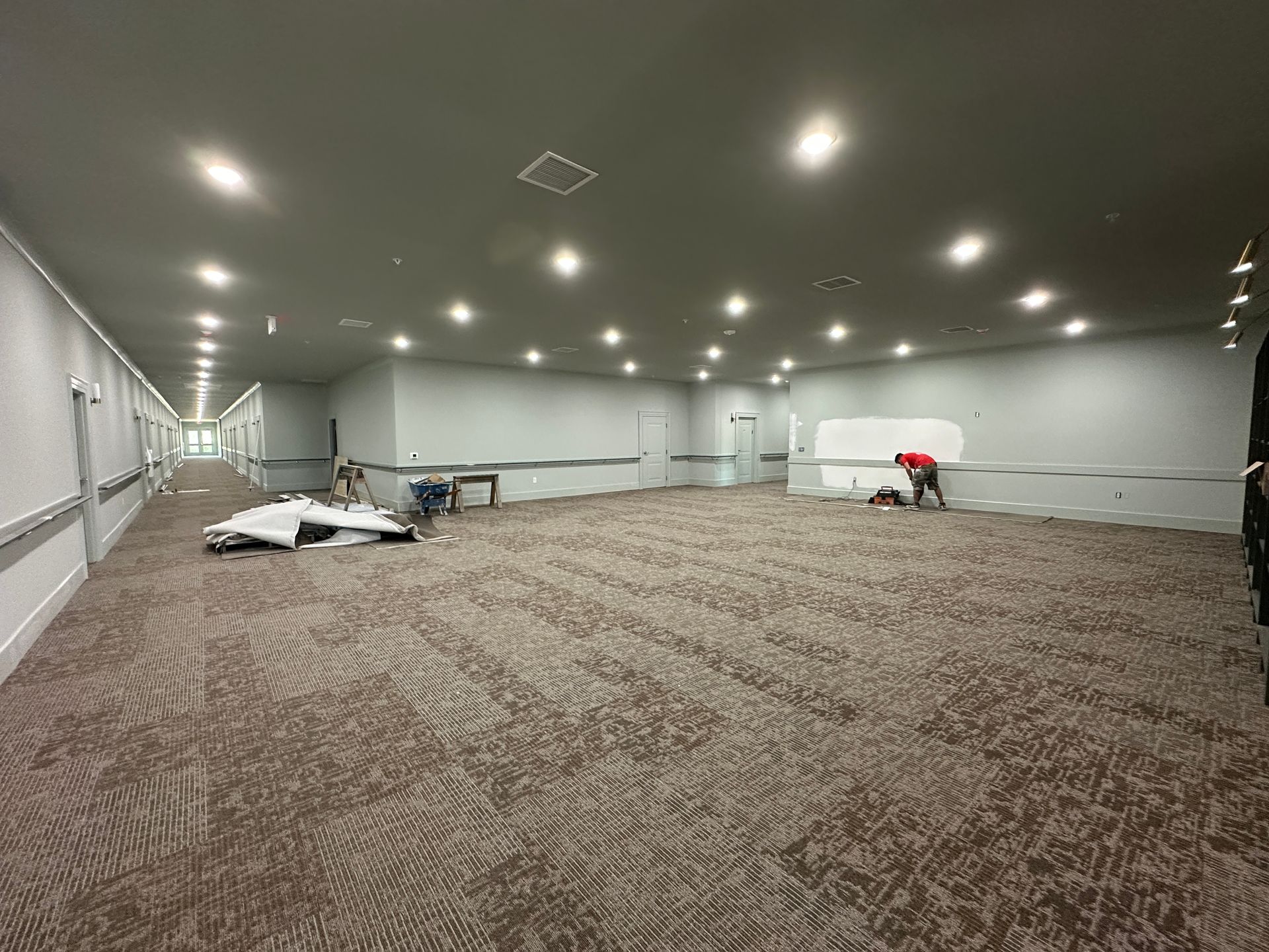 A large, empty event hall with grey walls, brown patterned carpet, recessed lighting, and workers installing trim.