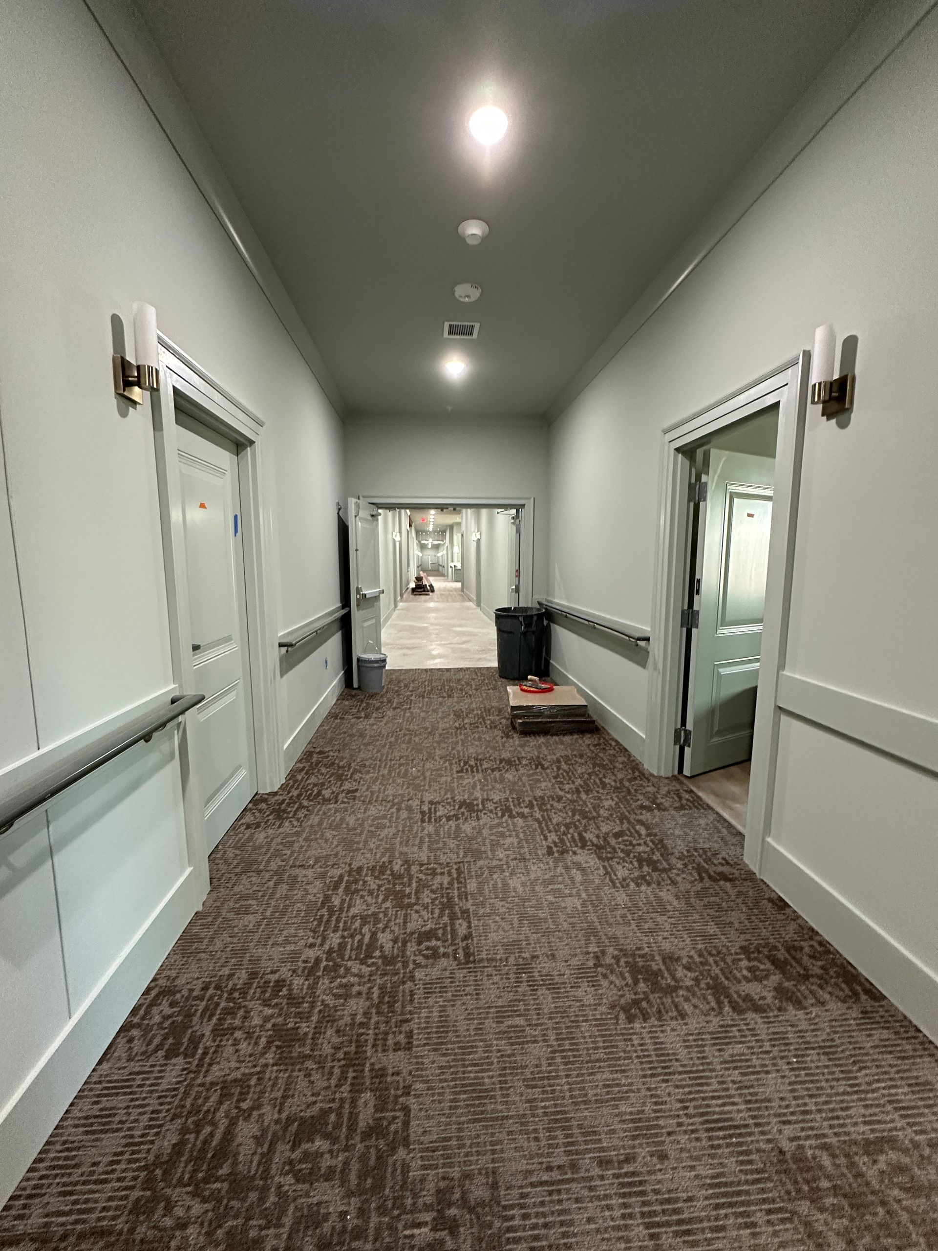 A narrow carpeted hallway with white walls and doors on both sides, leading to a brighter, open space in the background.