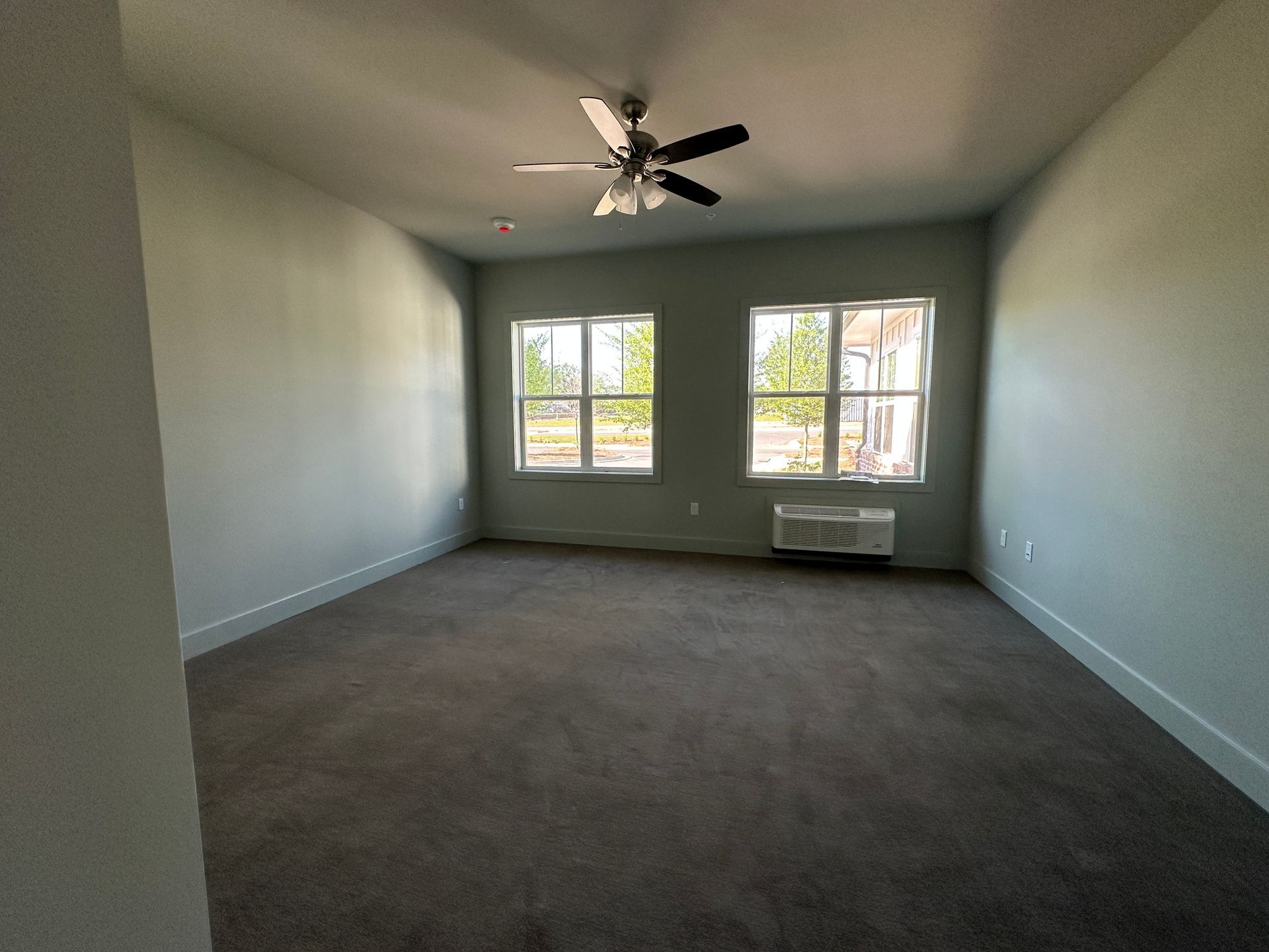 An empty room with light gray walls, dark carpeting, two windows, and a ceiling fan.
