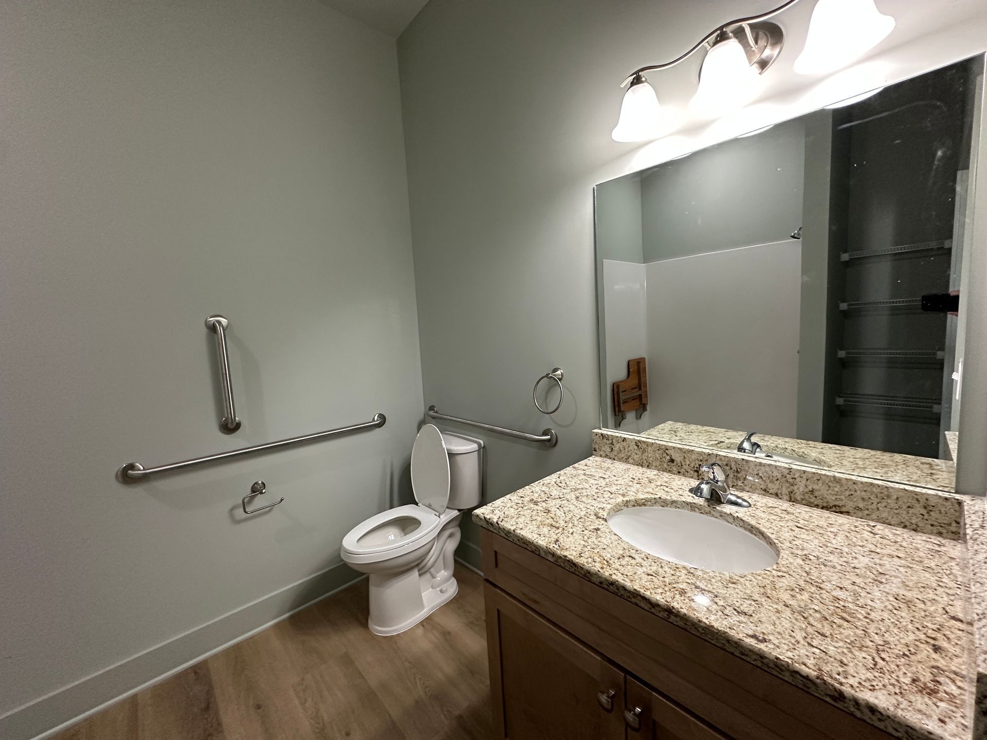An accessible bathroom with a granite-topped vanity, a toilet, and wall-mounted grab bars against light gray walls.