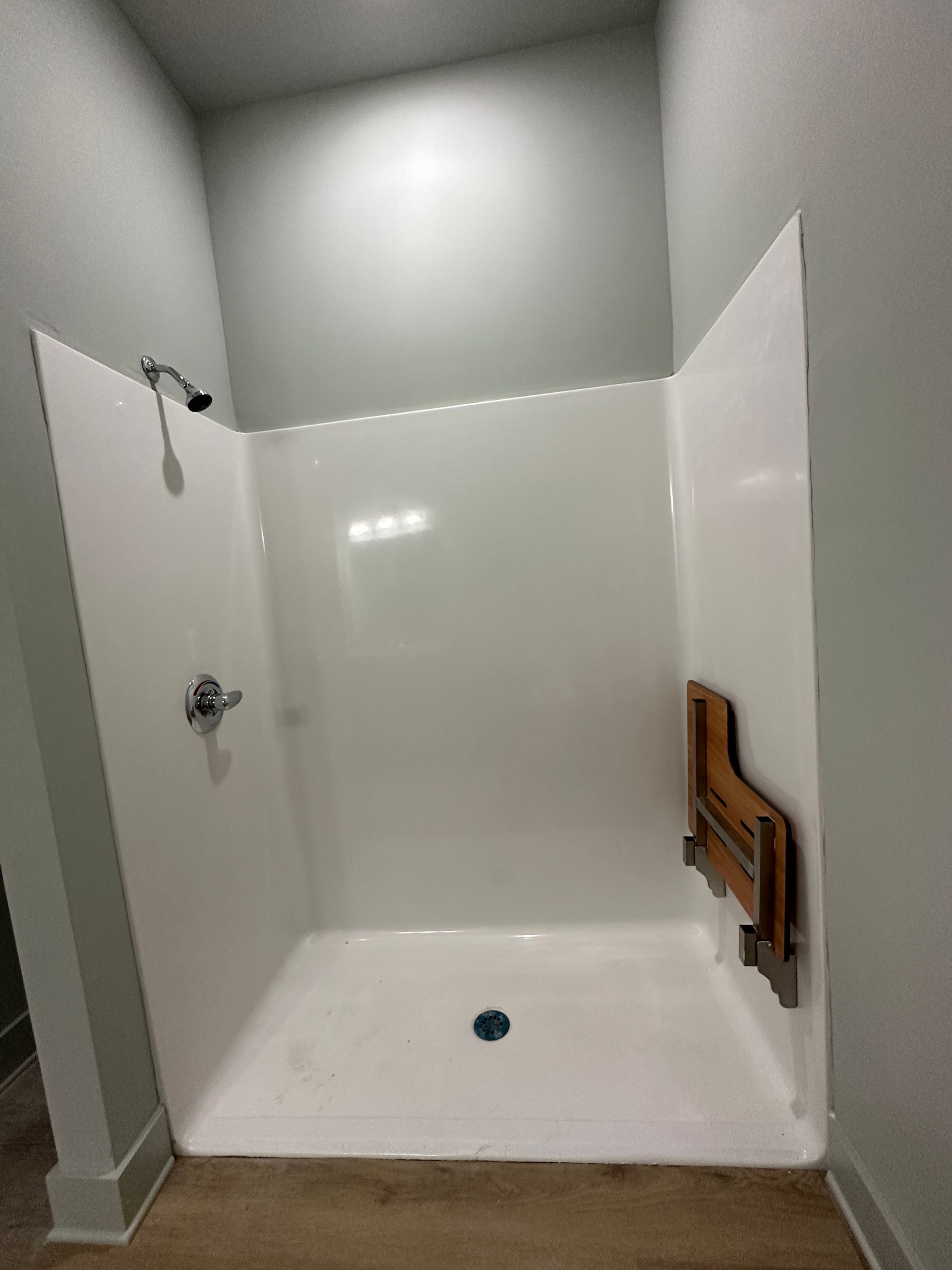 A walk-in shower with white walls, a chrome shower head, and a fold-down wooden seat mounted on the right side.