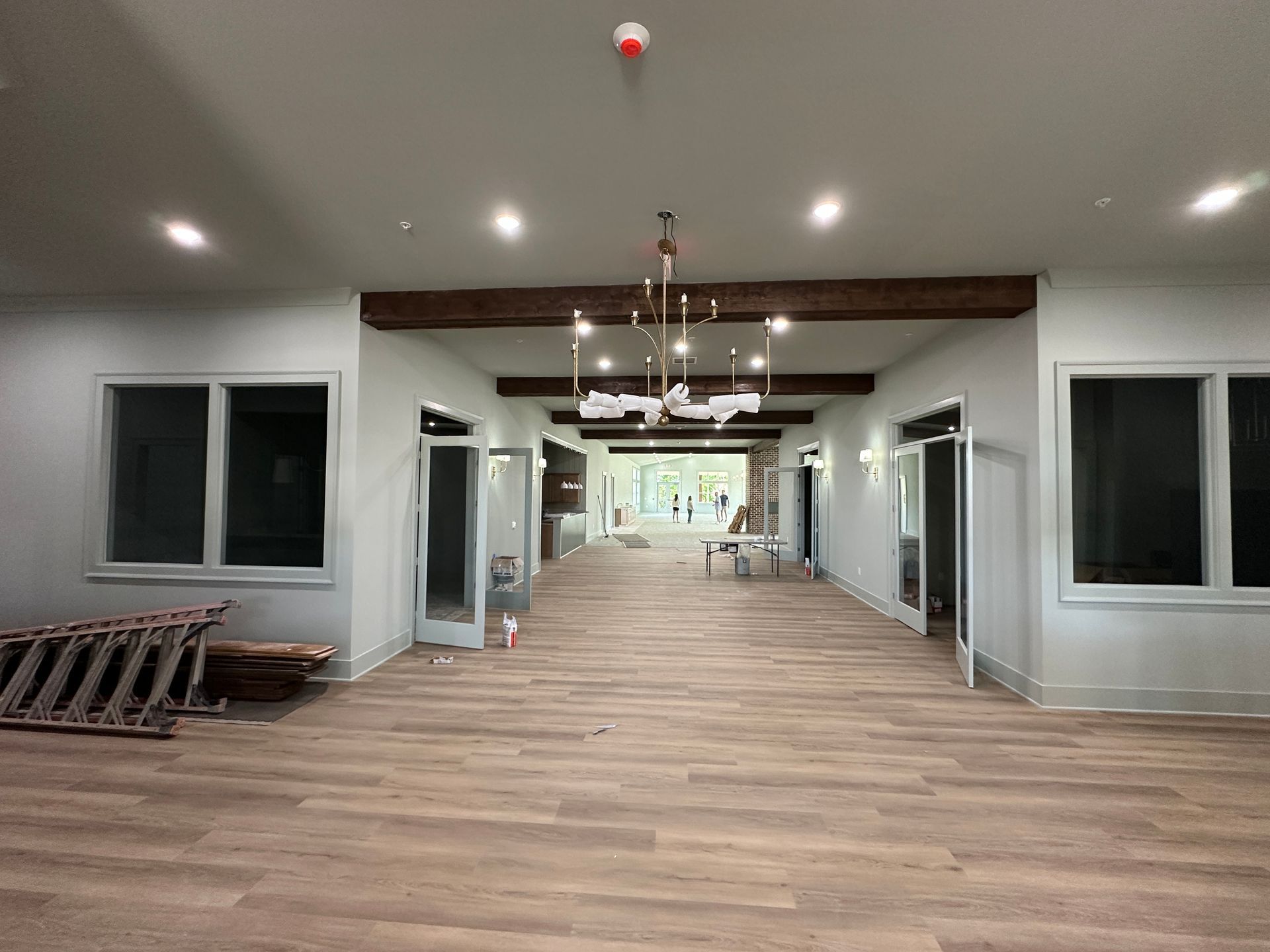 A long interior hallway with wood-style flooring, white walls, dark wooden ceiling beams, and an open doorway at the end.