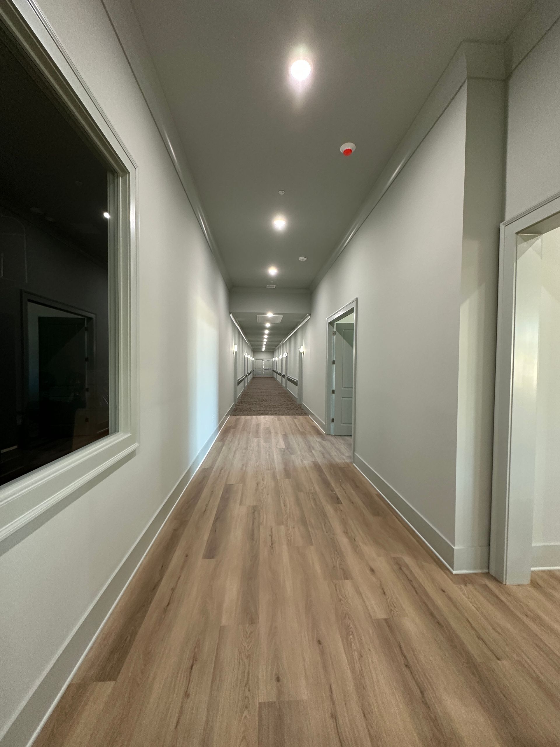 A long, brightly lit hallway with white walls and light wood-grain flooring, featuring doors on both sides.
