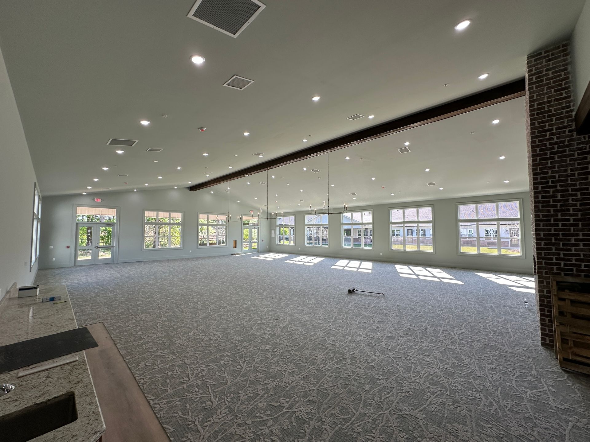 A spacious, empty event hall with grey patterned carpet, white walls, large windows, and recessed ceiling lighting.