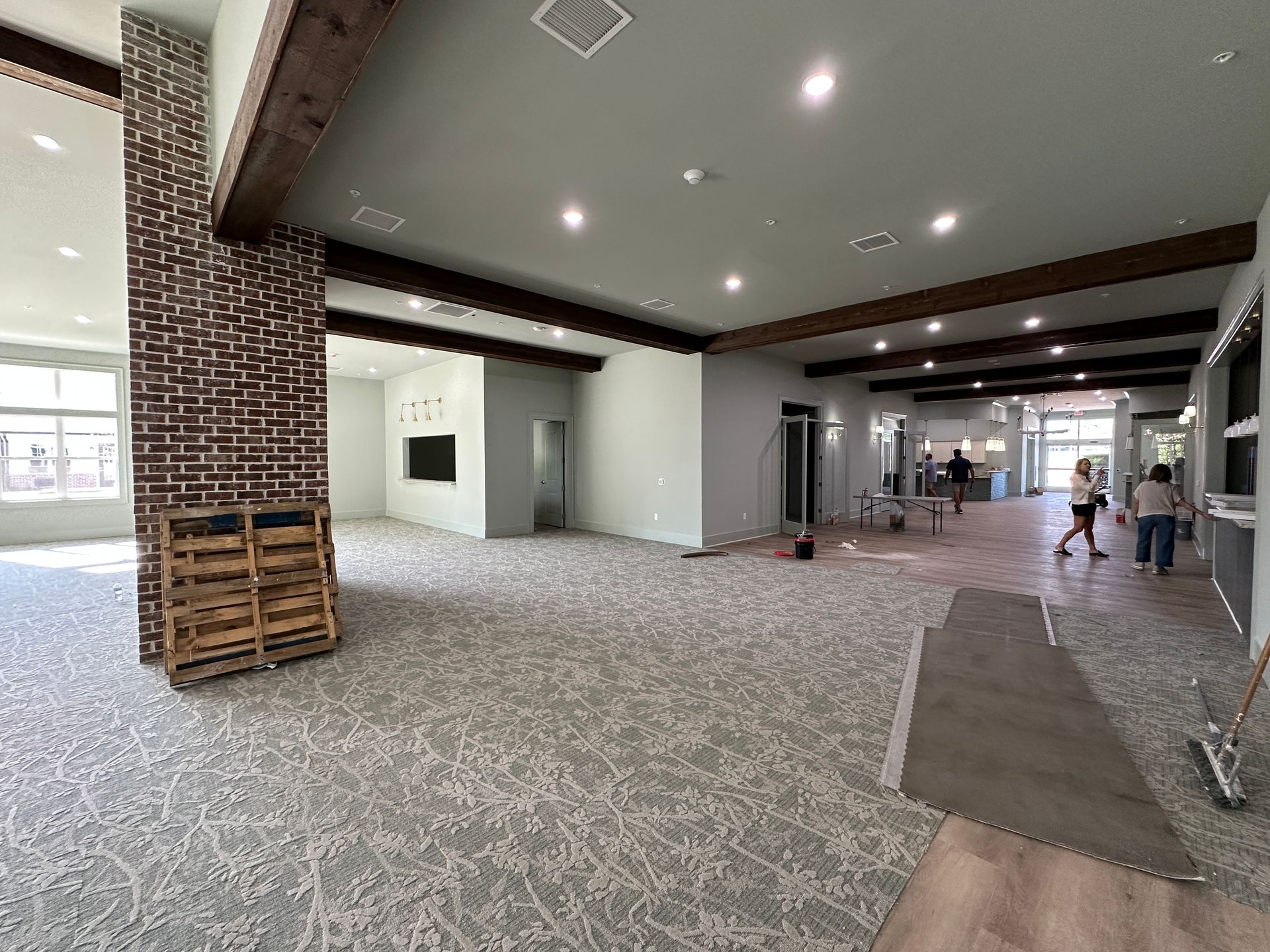 Spacious indoor lobby with exposed brick, dark wood ceiling beams, gray patterned carpet, and people walking in the back.