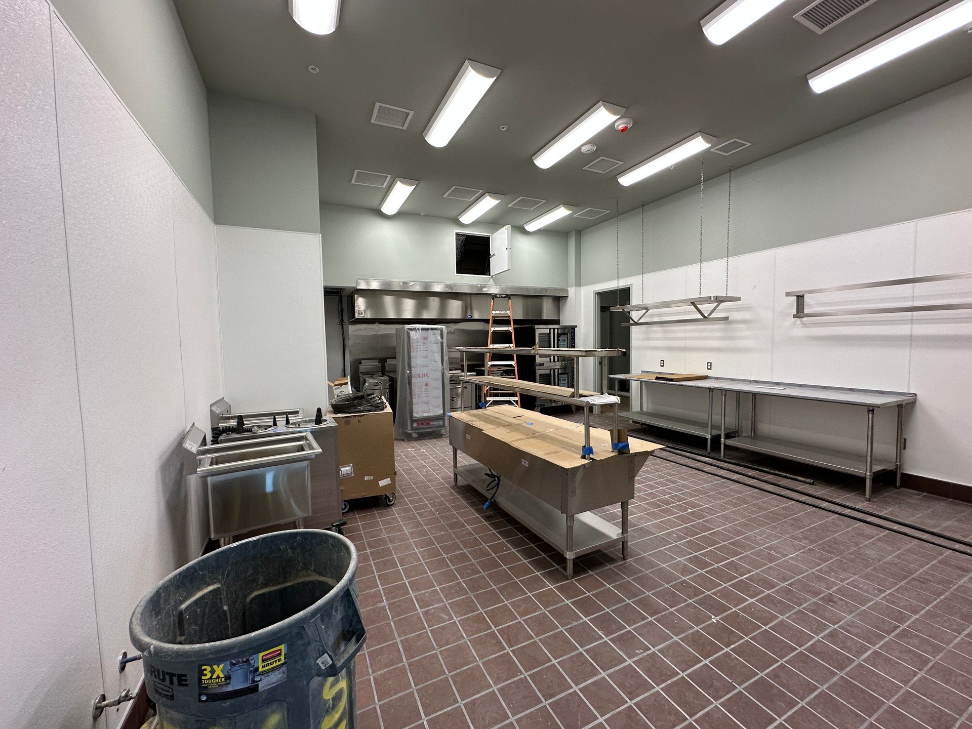 A commercial kitchen under construction with a central stainless steel workstation, tiled floor, and ceiling light fixtures.