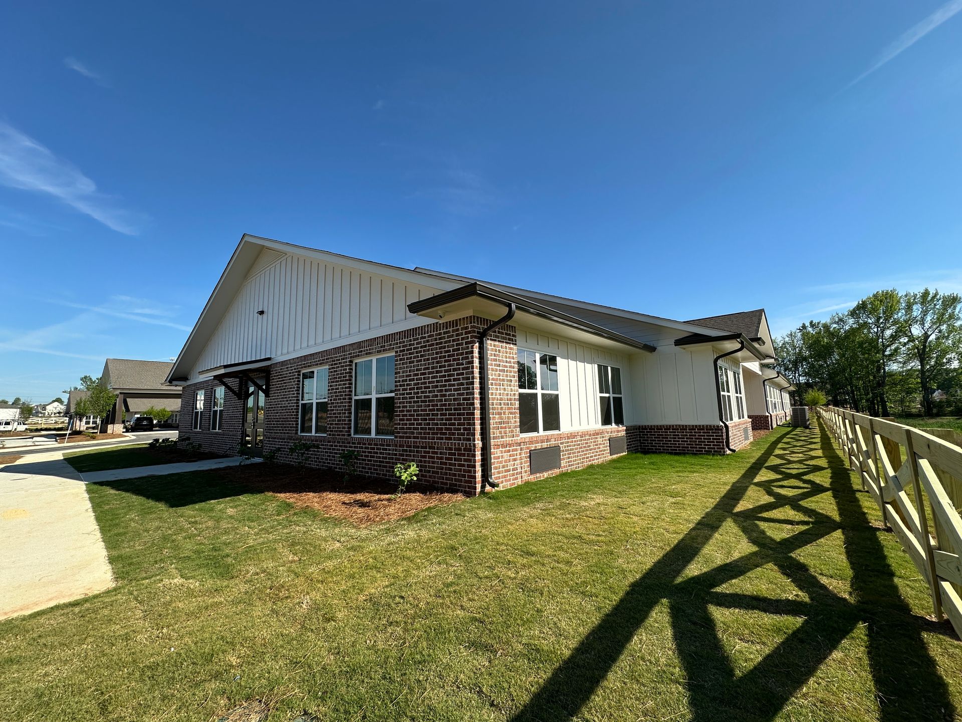 A one-story building with white siding and patterned brick accents, set in a grassy yard by a wooden fence under a blue sky.