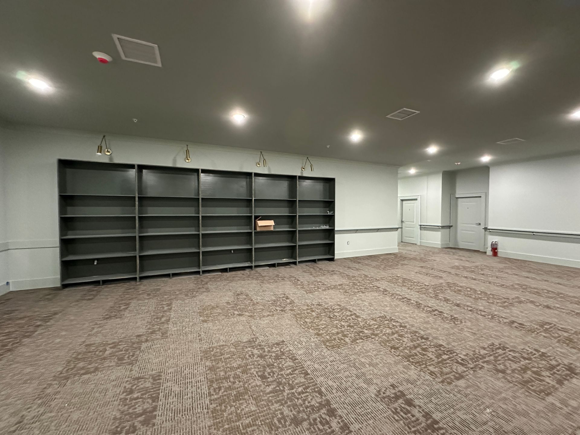 A wide, empty room featuring dark gray wall-to-wall shelving, patterned beige carpeting, and recessed ceiling lighting.