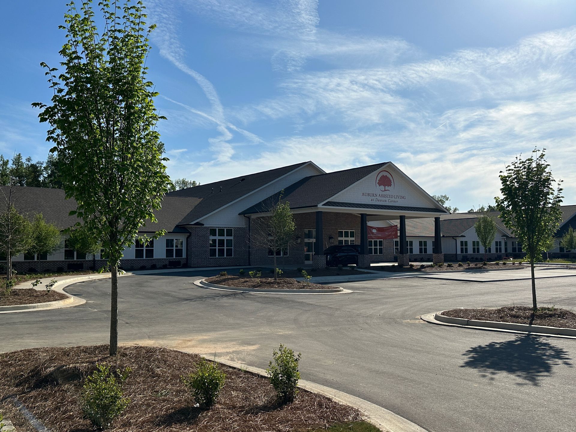 A single-story building with a covered entrance and stone accents, surrounded by a parking lot and young trees under a sky.