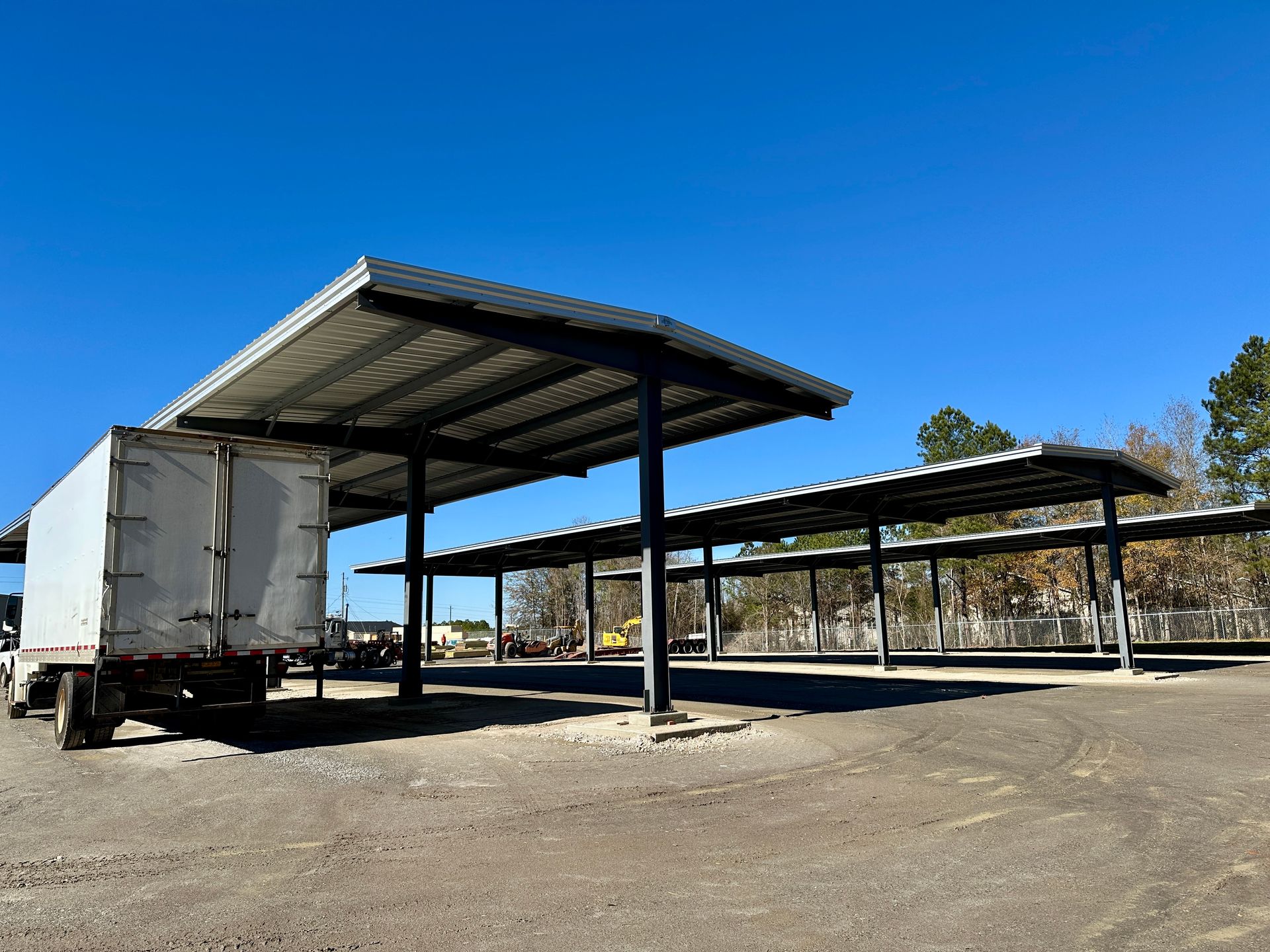 Semi-truck under a covered parking area with metal supports and roof; gravel lot, clear blue sky.