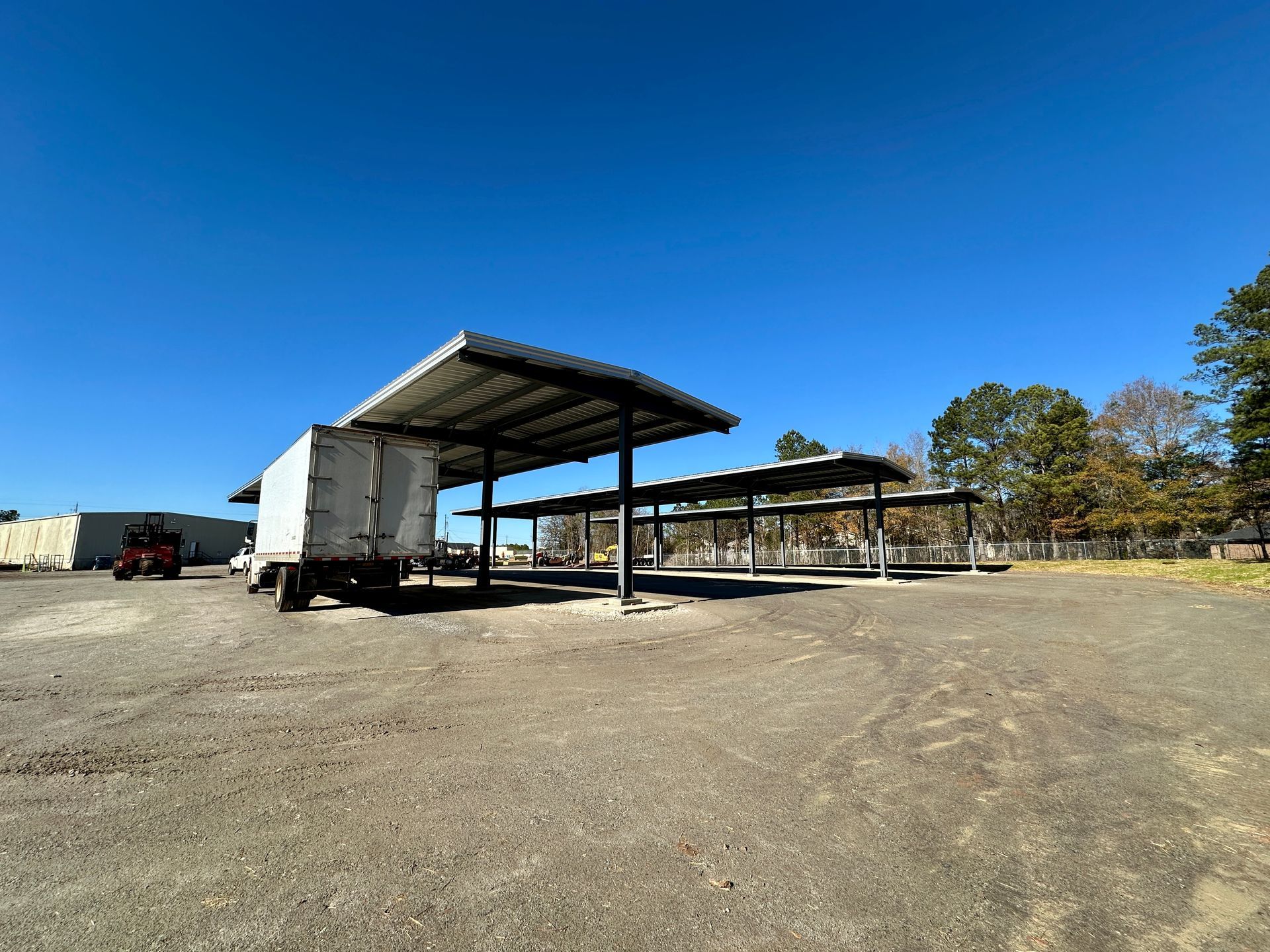 Empty gravel lot with a semi-trailer and covered shelters under a clear blue sky.