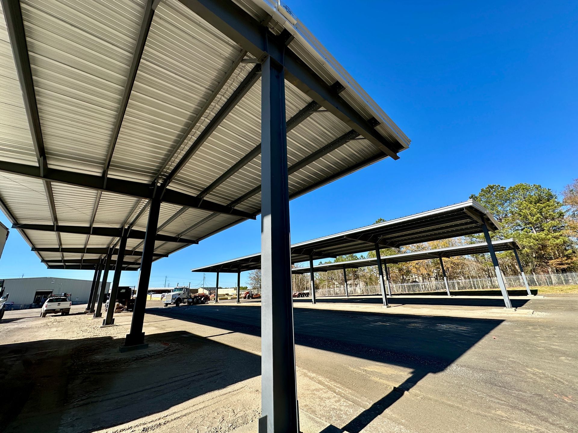 Metal carport structure on a sunny day. Gray roof, black support beams, and parked vehicles are visible.