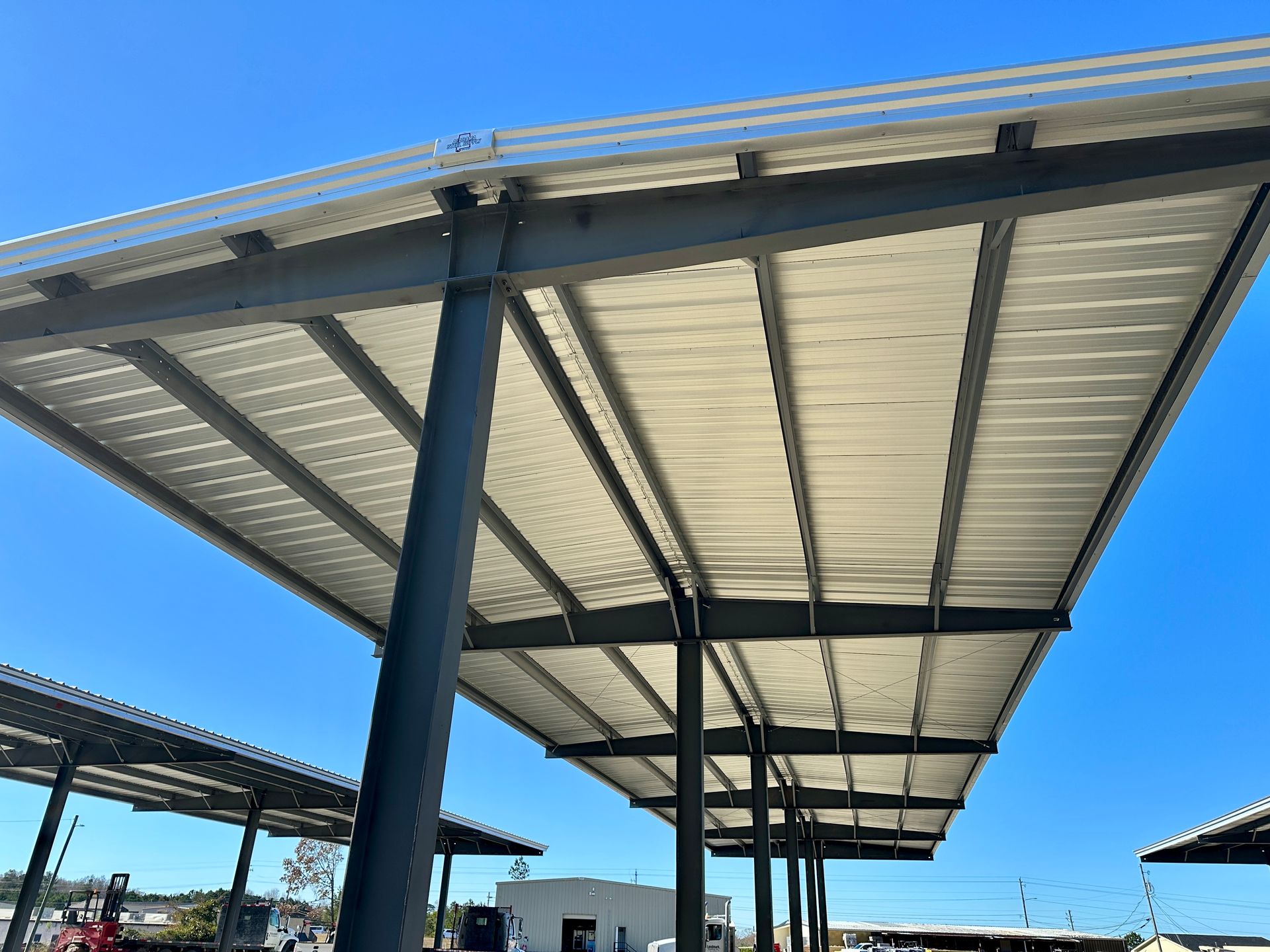 Metal carport with a corrugated metal roof, supported by dark steel beams and columns, against a blue sky.