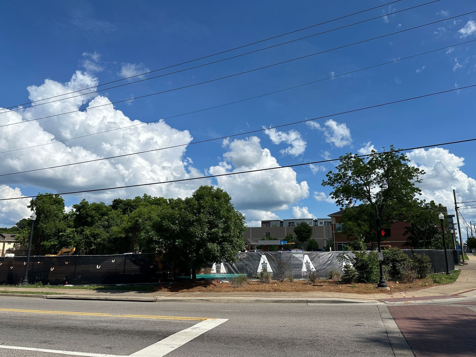 Street view of a fenced construction lot with trees, buildings, and a blue sky with clouds