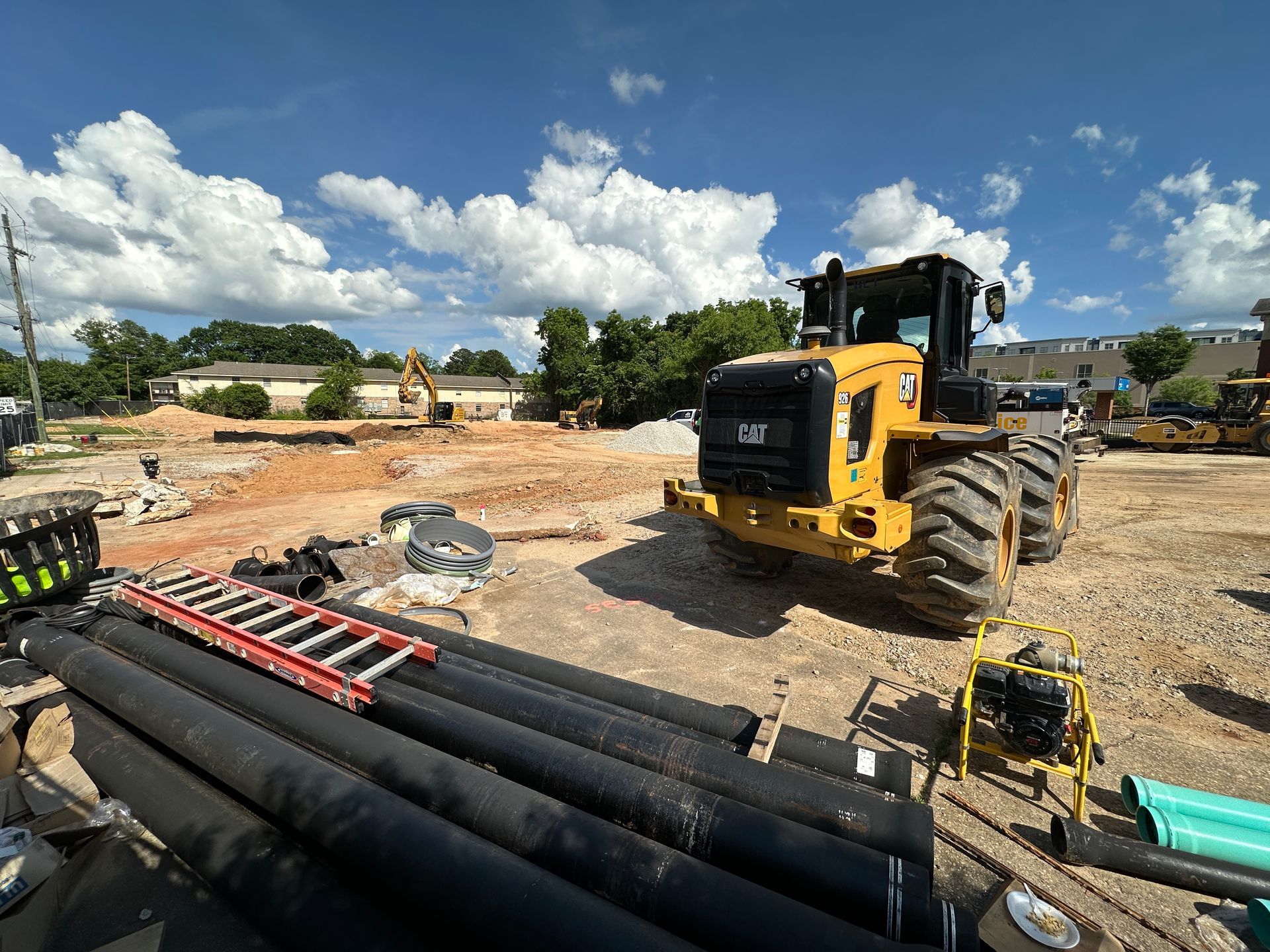 Yellow construction vehicle at a building site with pipes and equipment under a cloudy blue sky