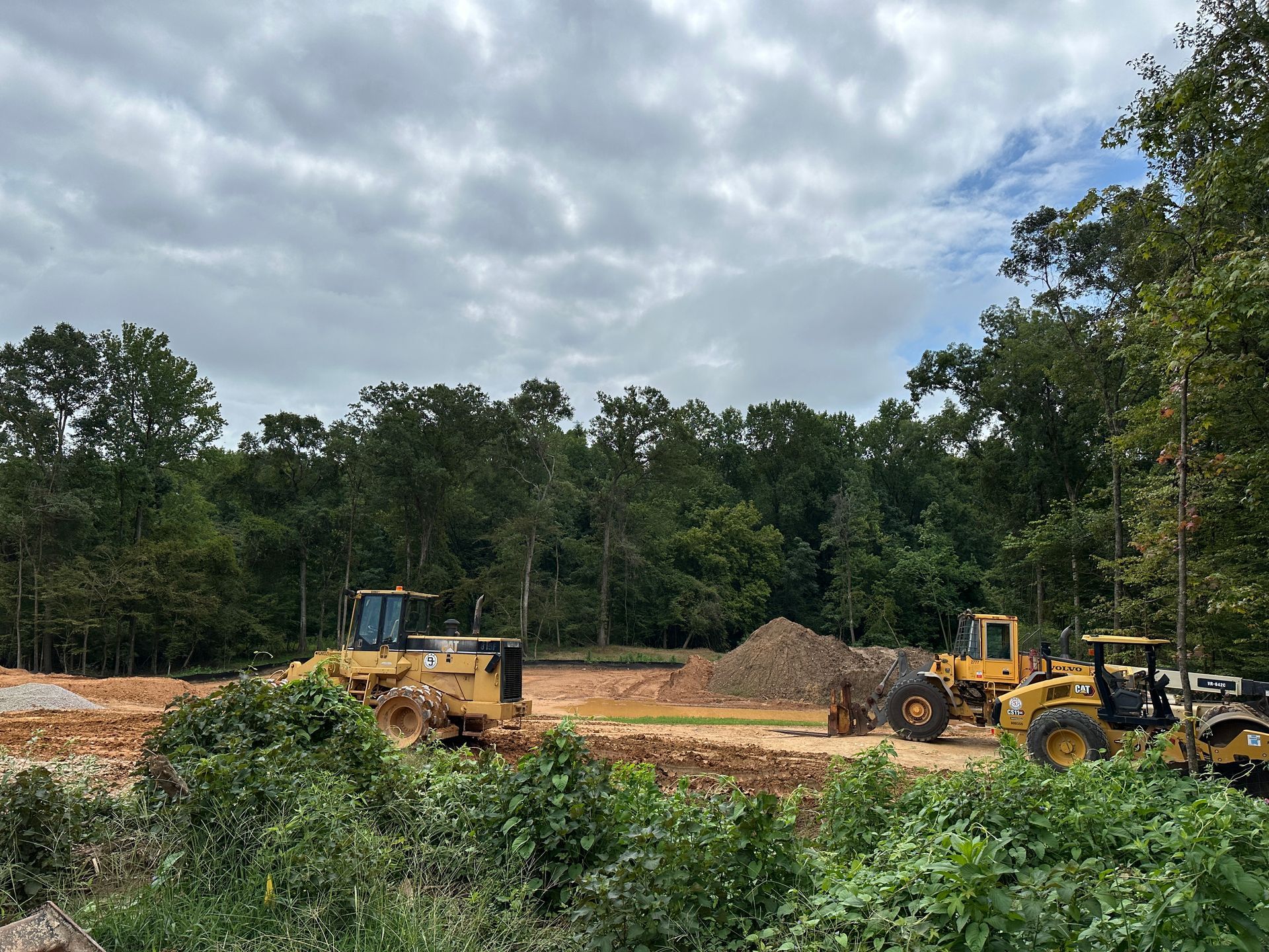 Construction site with yellow bulldozers and equipment surrounded by trees under a cloudy sky.