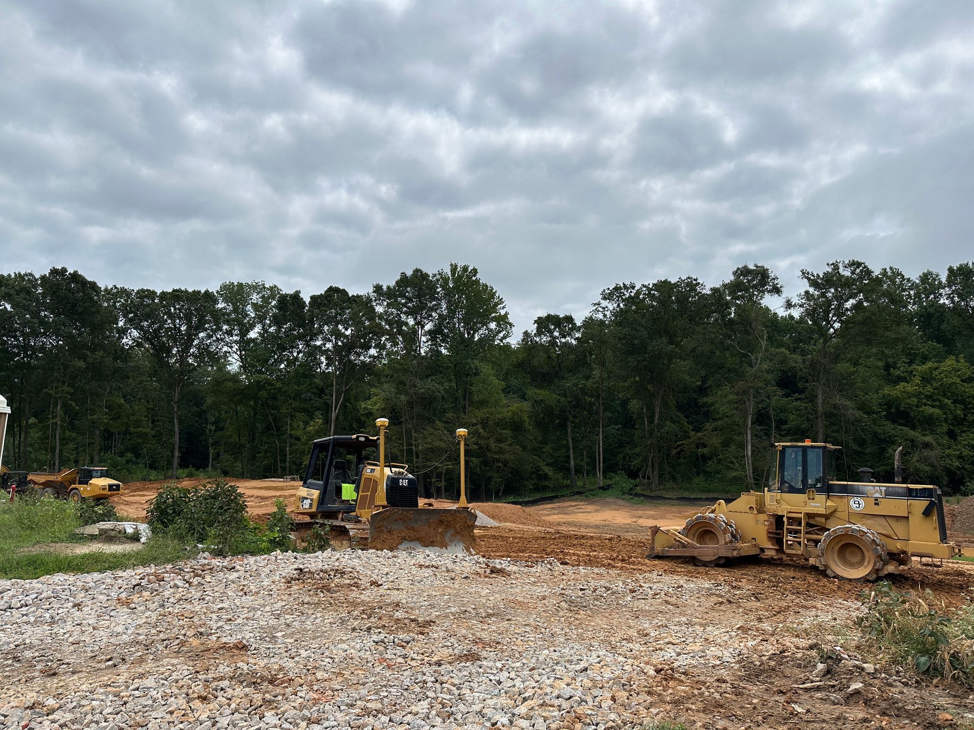 Two bulldozers on a construction site clearing land with trees in the background under a cloudy sky.