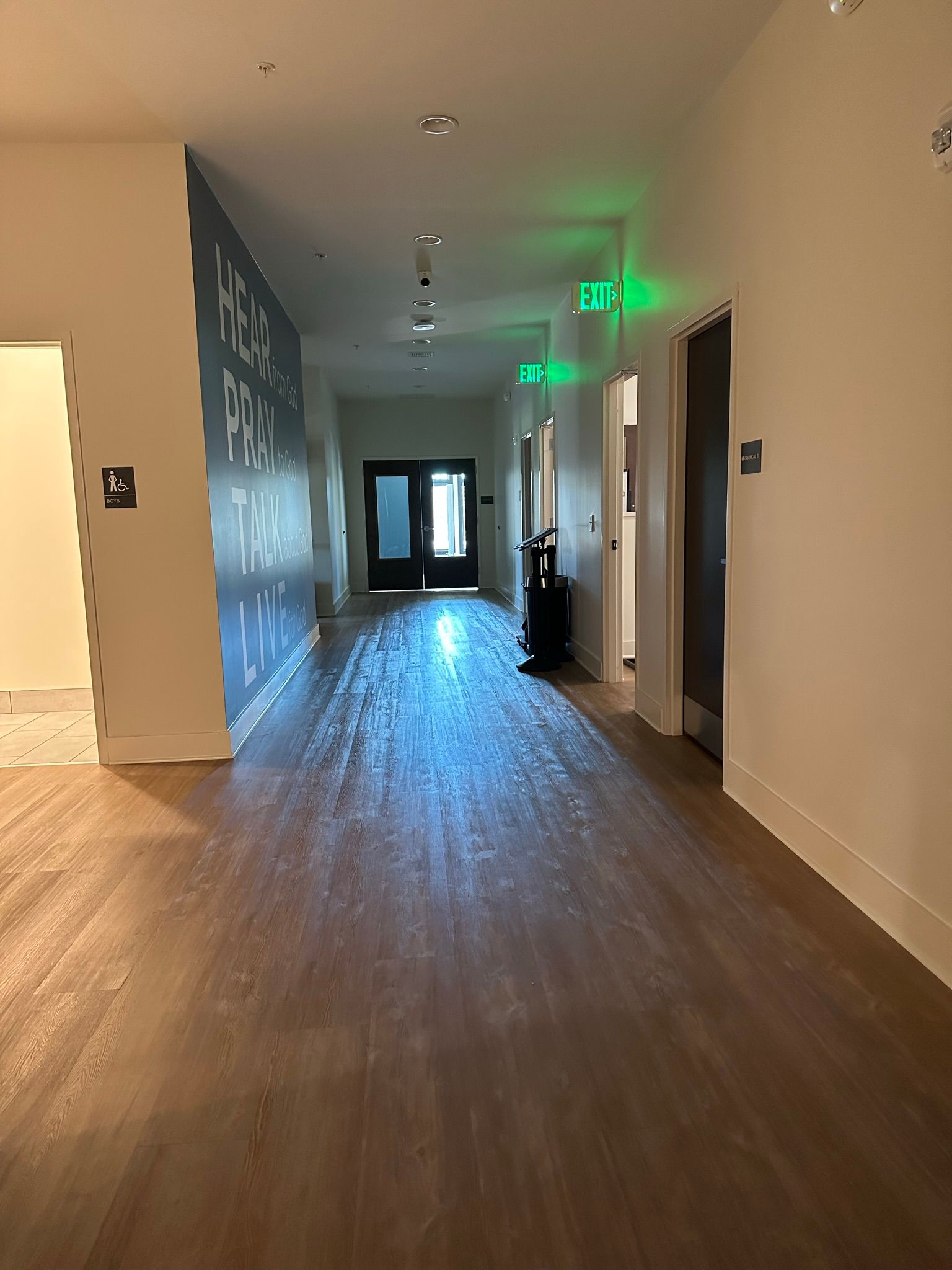 A long hallway with wooden floors and a green exit sign.
