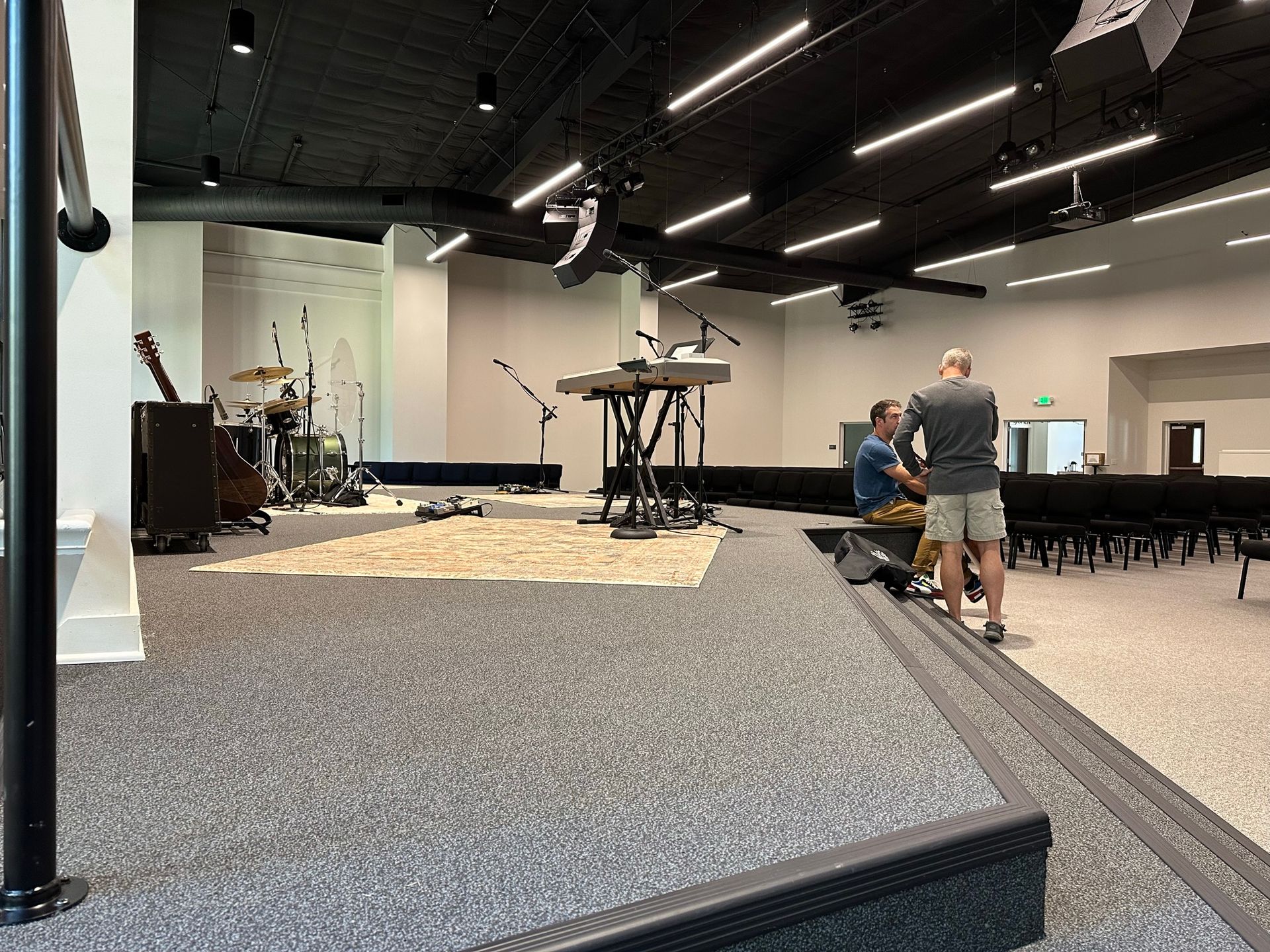 Two men are standing in a large room with a piano and a drum set.