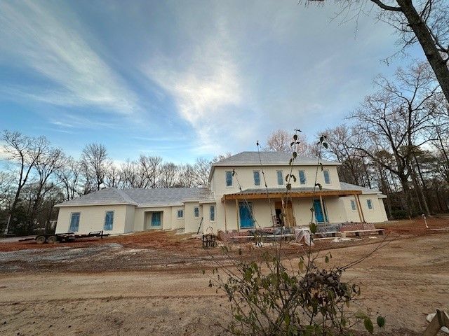 Large cream-colored house under construction, blue sky background. Bare trees surround the house.