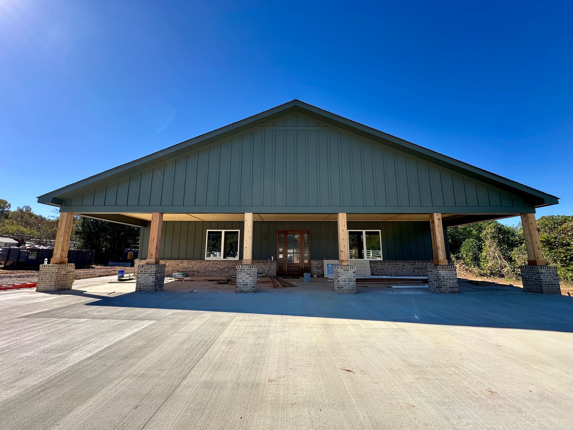 Building under construction; green siding, brown support beams, two windows, concrete driveway, clear blue sky.