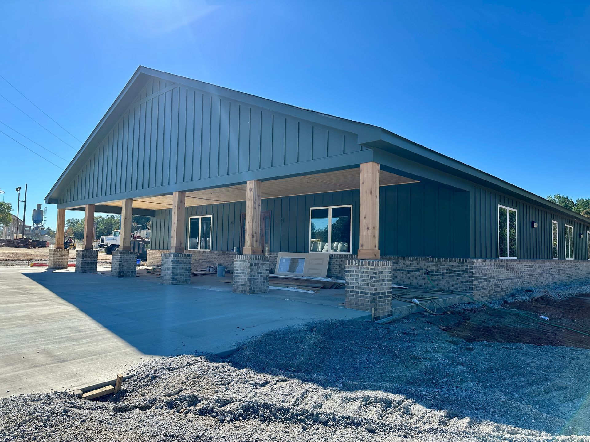 A building under construction with green siding, a covered porch, and stone columns on a sunny day.
