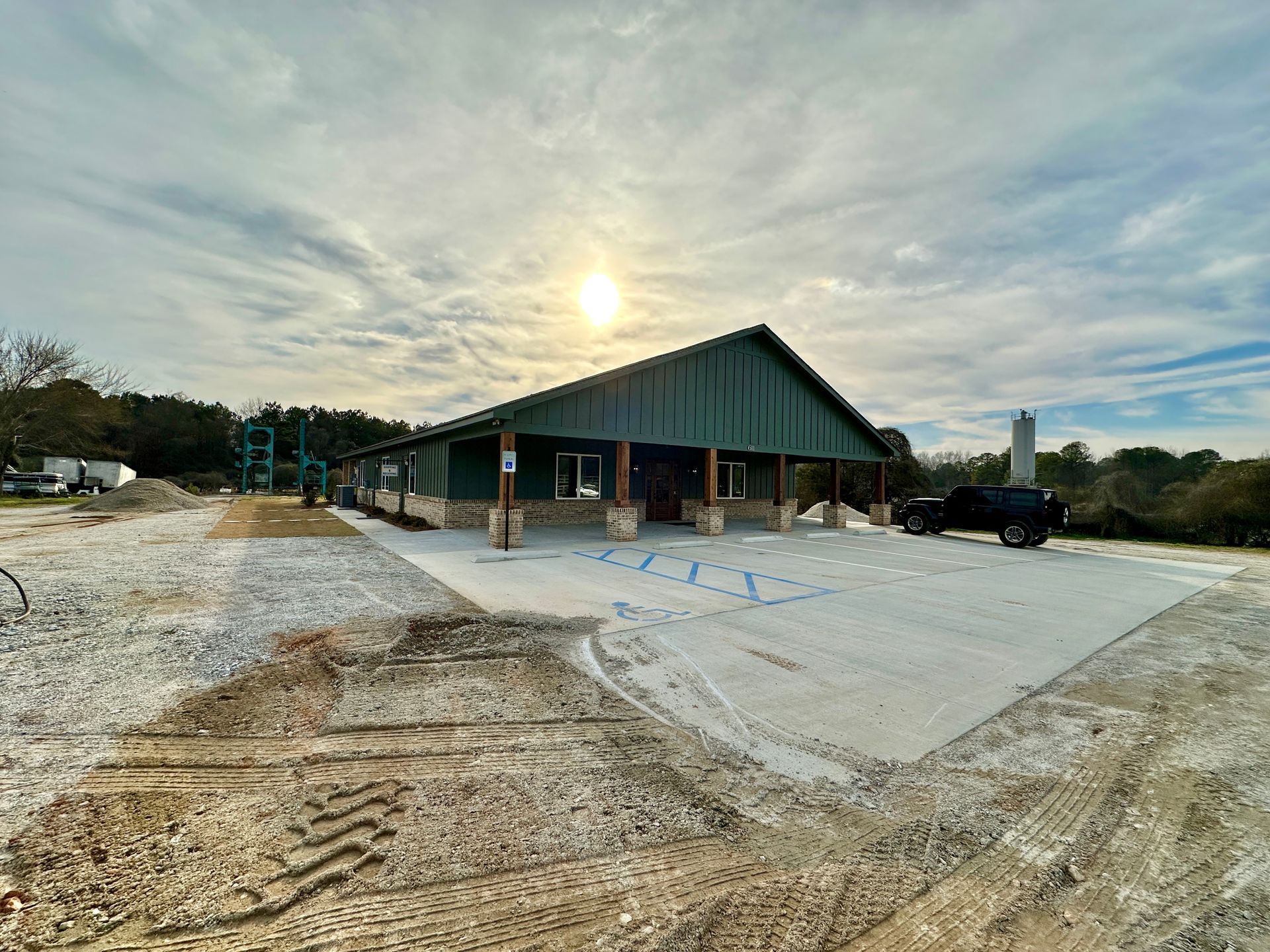 Building under construction with a large concrete parking area. Overcast sky with sun, dirt and gravel.