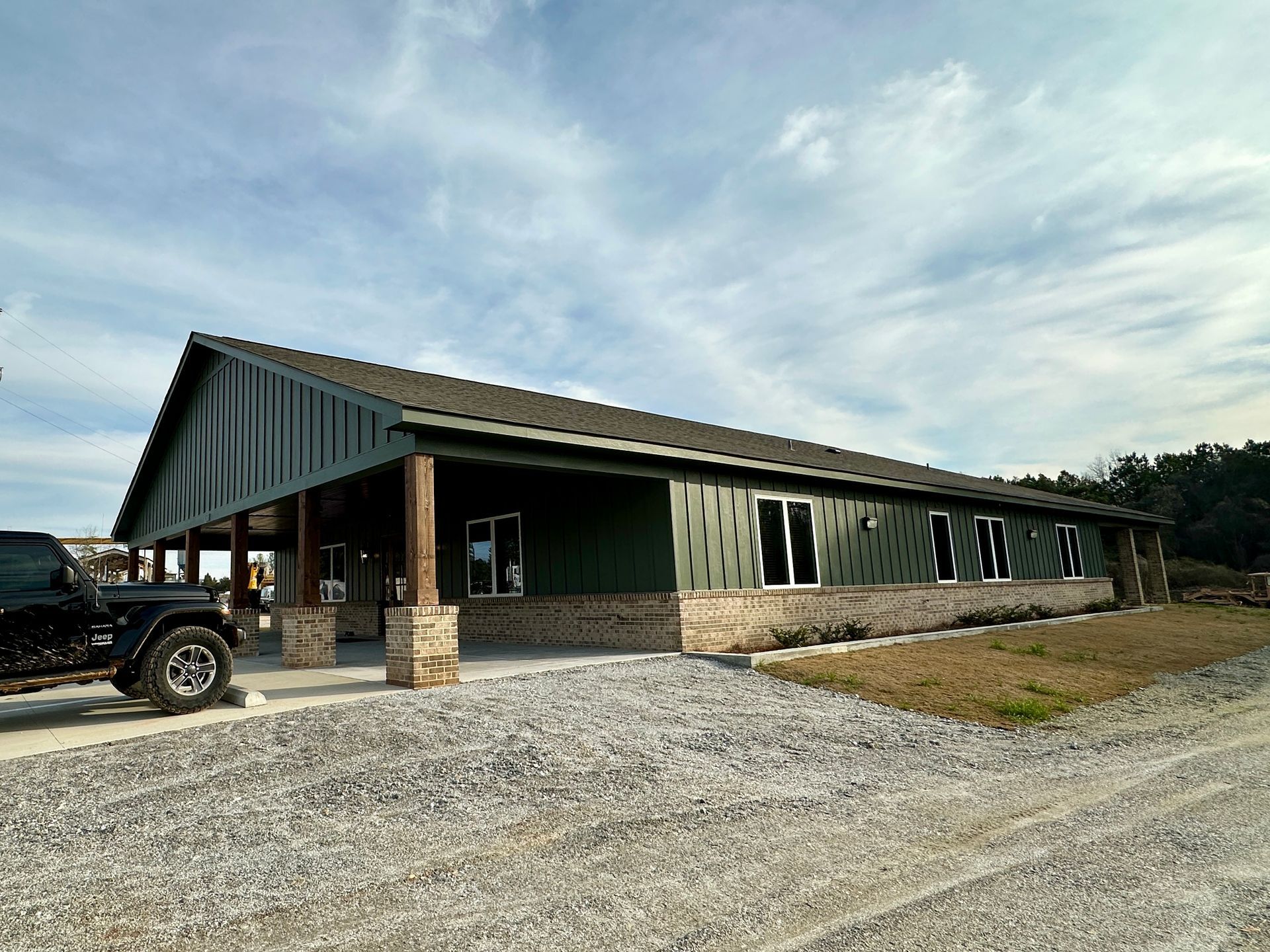 Green building with stone accents, covered porch, gravel driveway, overcast sky.