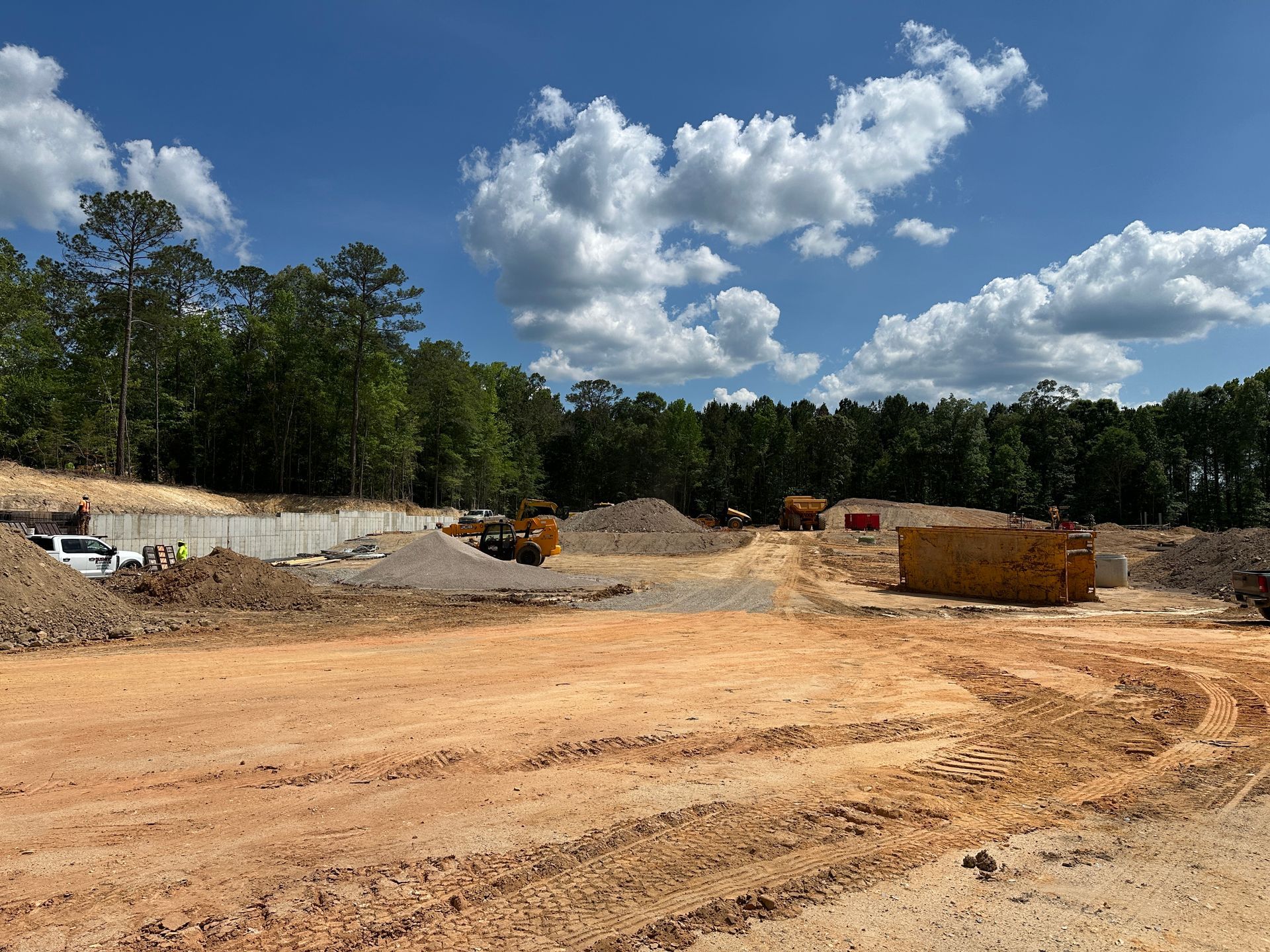 Construction site with dirt piles, heavy equipment, and a tree line under a partly cloudy blue sky