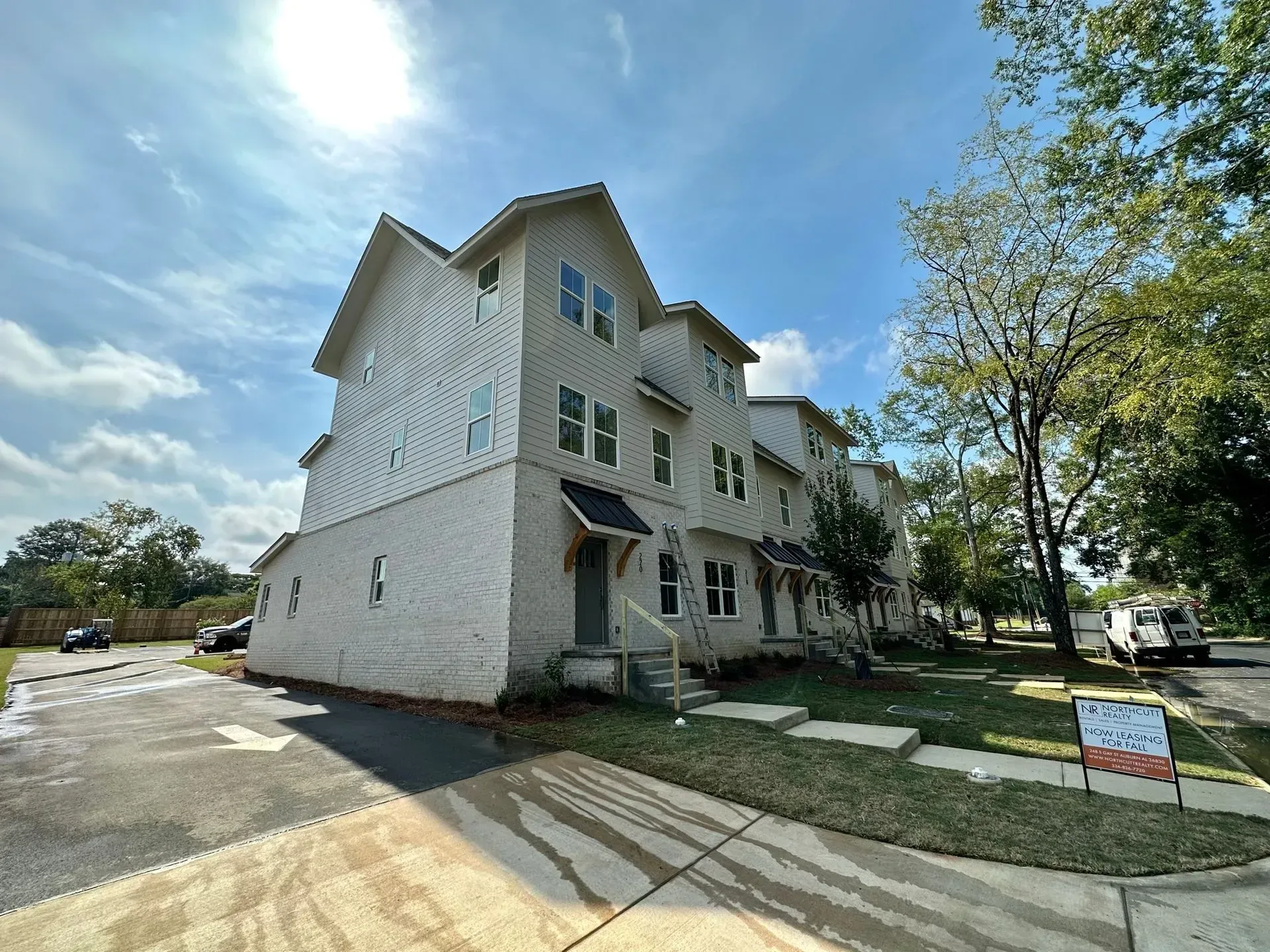 Three-story white multi-family townhome with a front lawn, driveway, and a small sign under a blue sky.