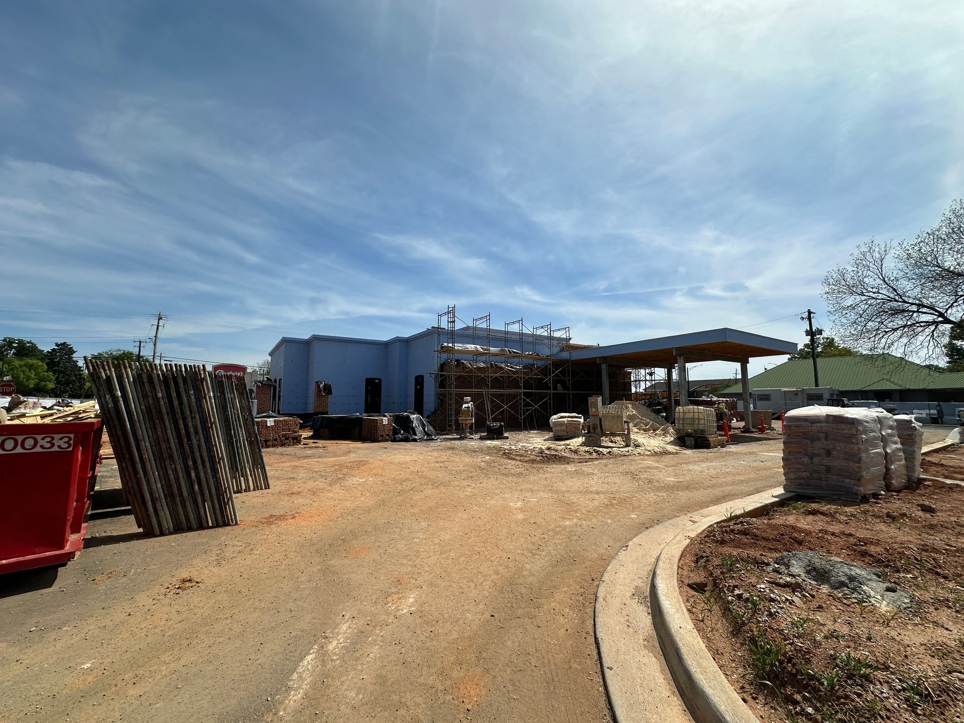A construction site with an unfinished building, a covered driveway, building materials, and a red dumpster under blue sky.