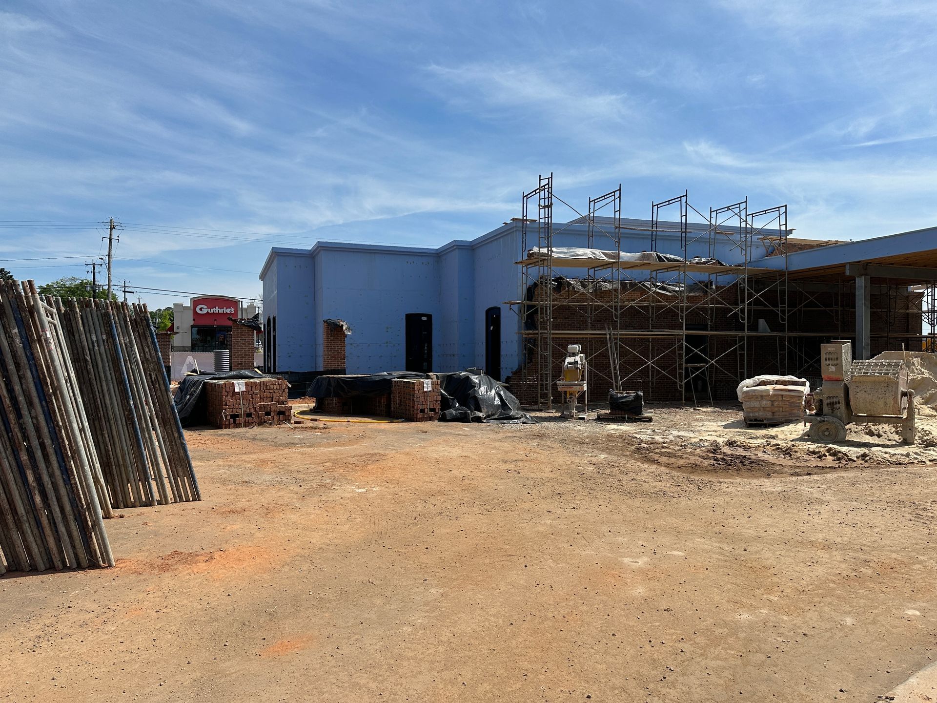 Construction site with a light blue building, scaffolding, stacked pallets, and a cement mixer under a sunny blue sky.