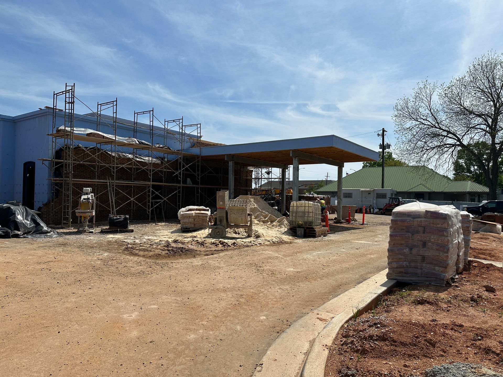 A construction site with a partially blue building, scaffolding, and stacked bags of materials on a gravel lot.