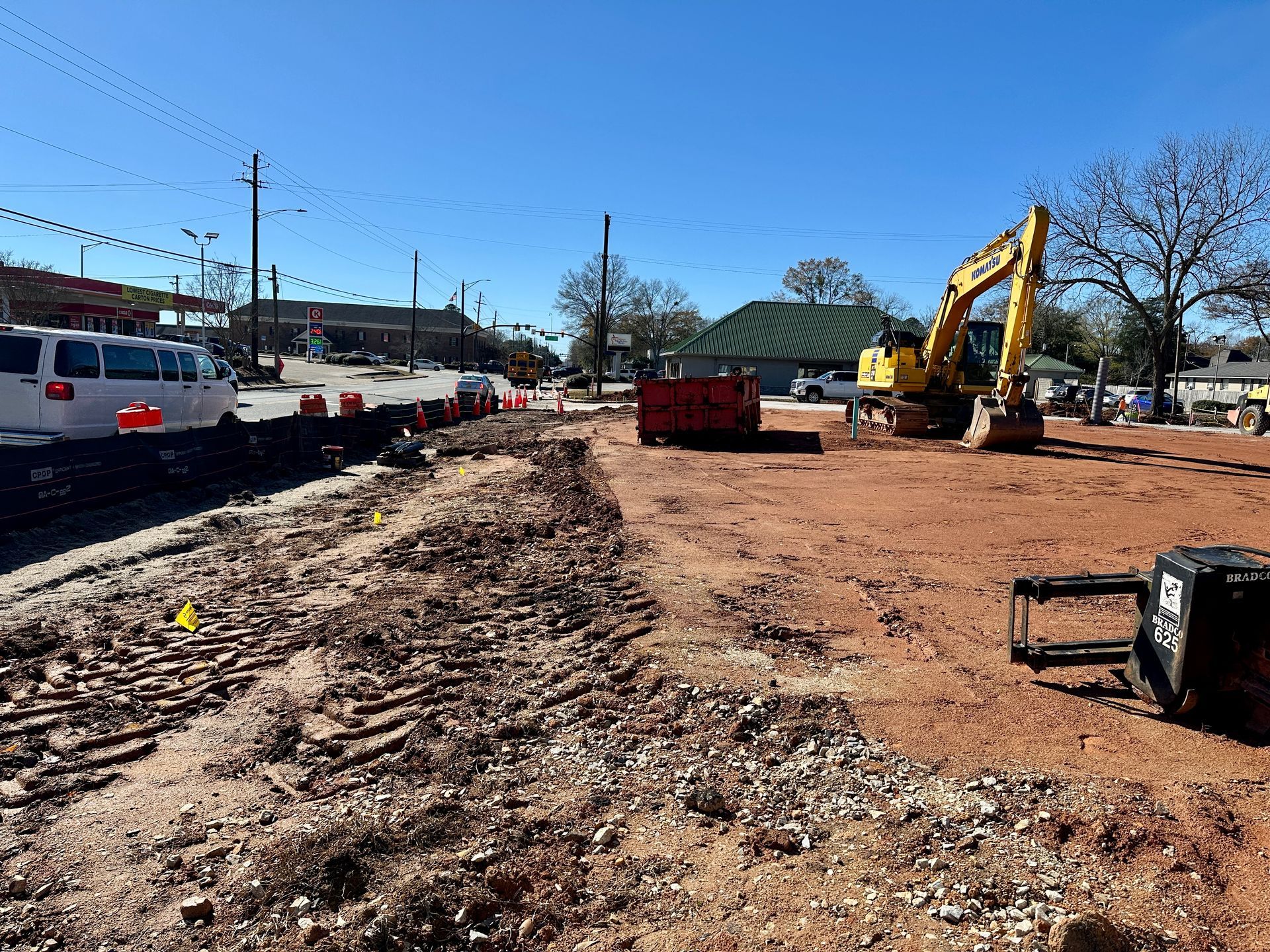 Construction site with excavator and red dirt, next to a street with cars and buildings.