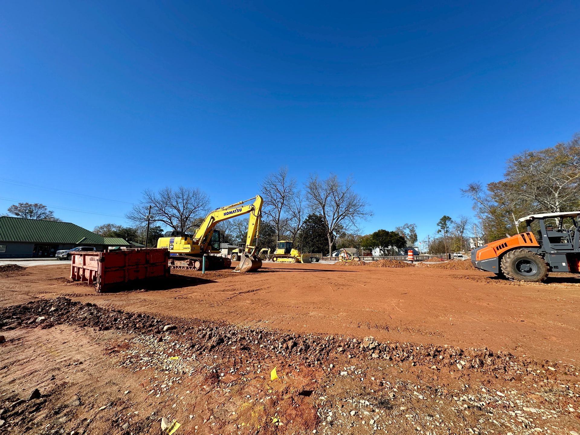 Construction site with excavator and roller on red dirt under a blue sky.
