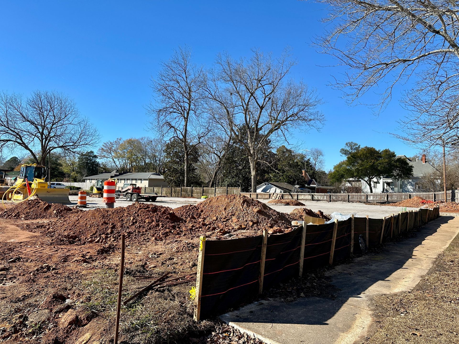 Construction site with dirt piles, fencing, and trees against a blue sky.