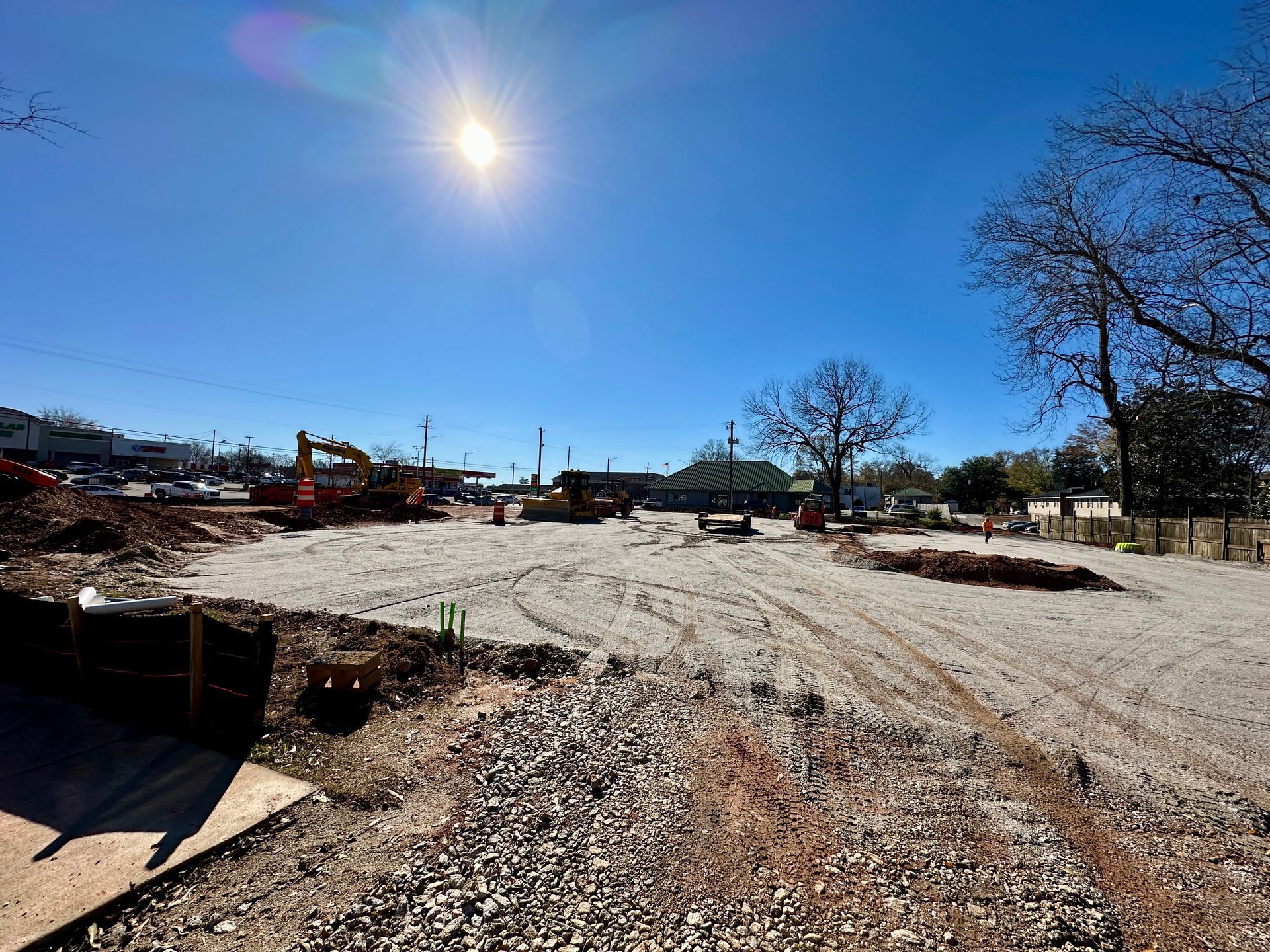 Construction site under a bright sun. Gravel covers the cleared ground, with heavy machinery visible.