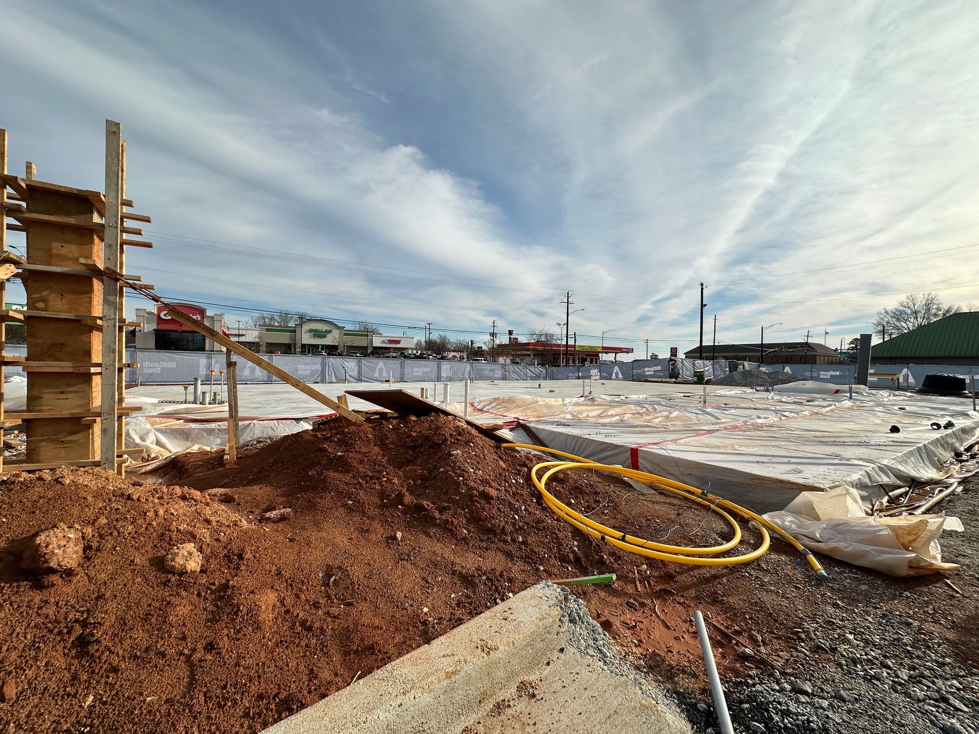 Construction site with exposed foundation, dirt piles, and wooden structures against a cloudy sky.