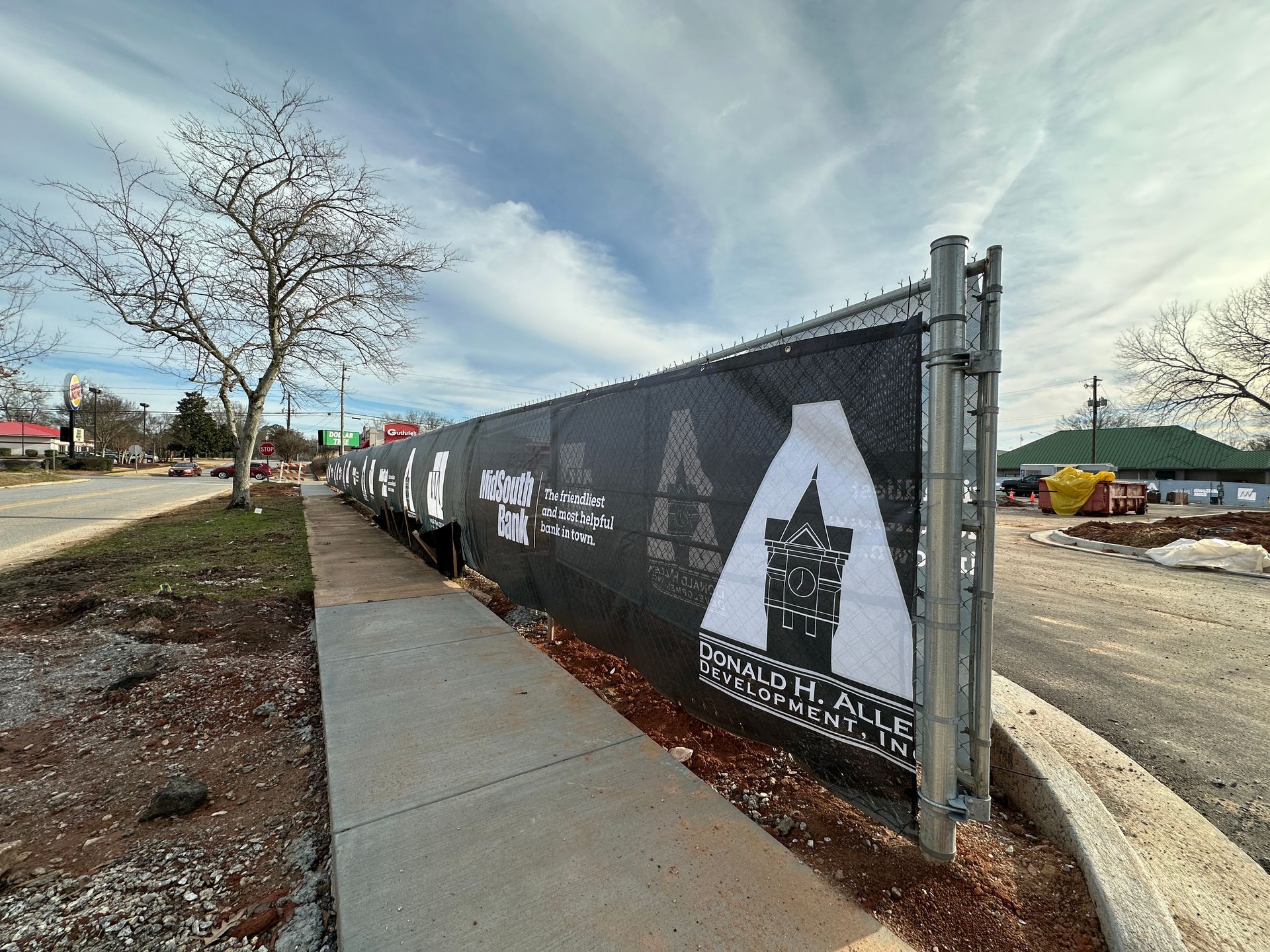 Construction site behind a black fence with a banner. Gray sidewalk along a road and a cloudy sky above.