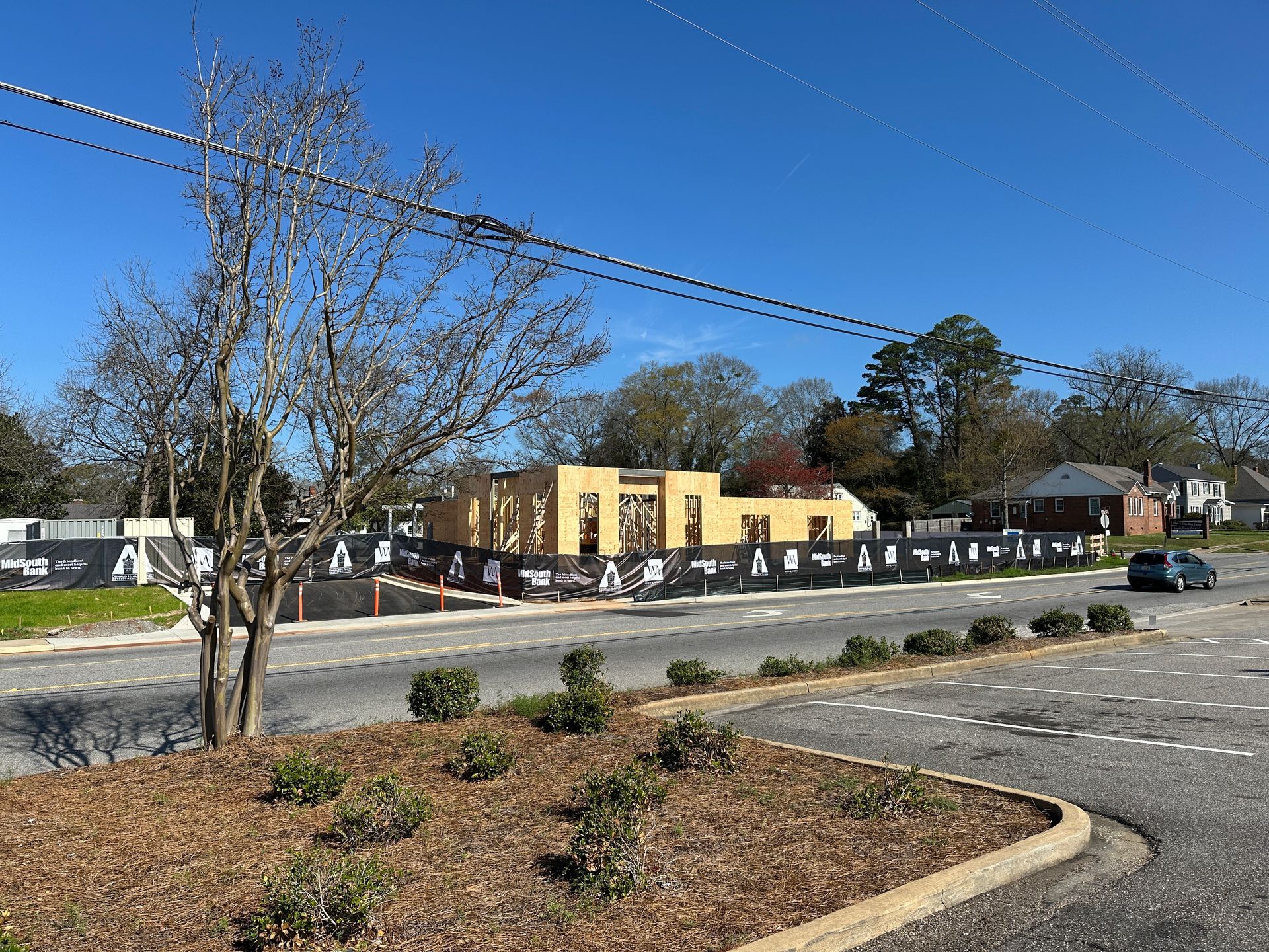 A wood-framed building under construction, surrounded by a black privacy fence, sits next to a paved road and parking lot.