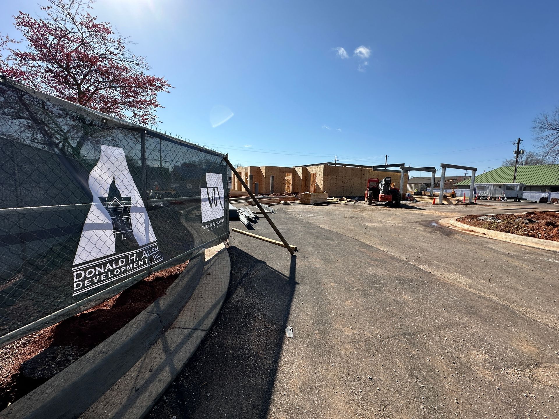 Construction site with wooden framing, a black mesh privacy fence with a logo, and a blue sky in the background.