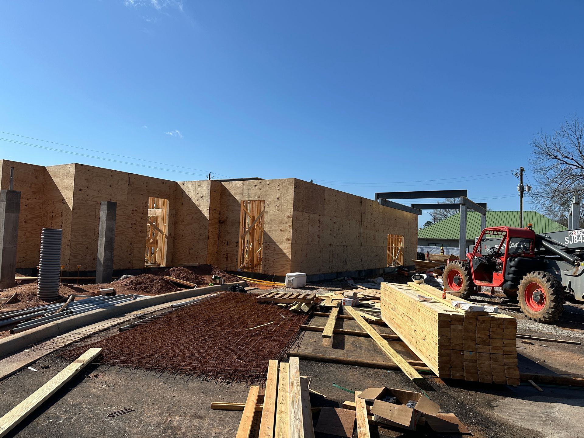 A construction site with a partially framed wooden building, construction materials, and a red telehandler under a blue sky.